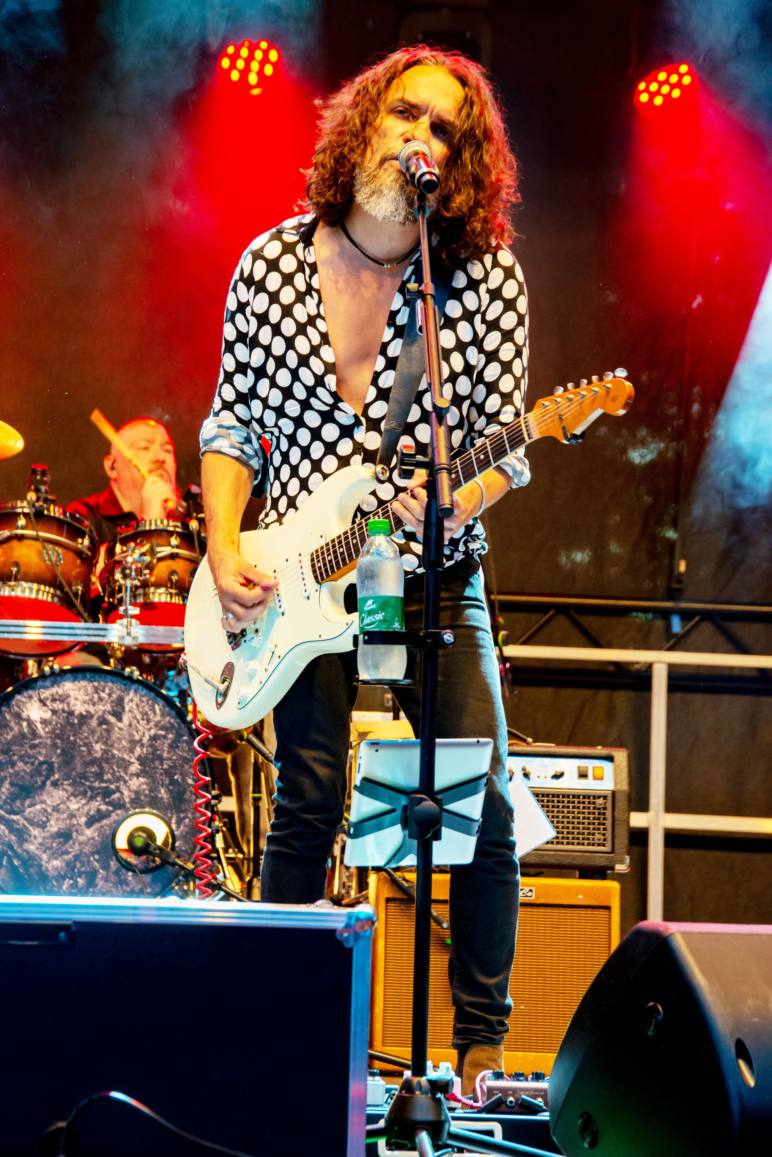 Male musician with curly hair and beard playing an electric guitar on stage, wearing a black and white polka dot shirt, with a drummer in the background.