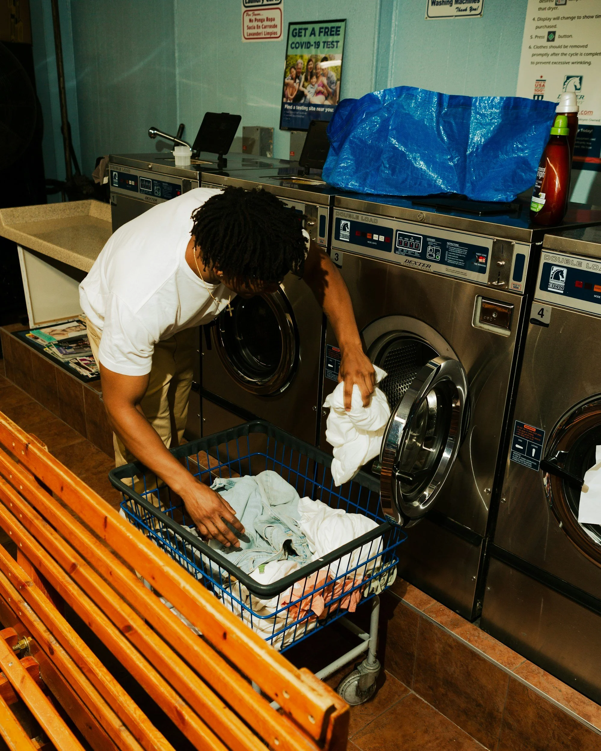 A man with dreadlocks wearing a white shirt is loading clothes into a washing machine in a laundromat. There is a blue shopping bag on top of the washing machines and a bottle of laundry detergent. A cart with clothes is in front of the man, and magazines are on a shelf nearby. Signs about COVID-19 testing and laundry instructions are visible on the wall.