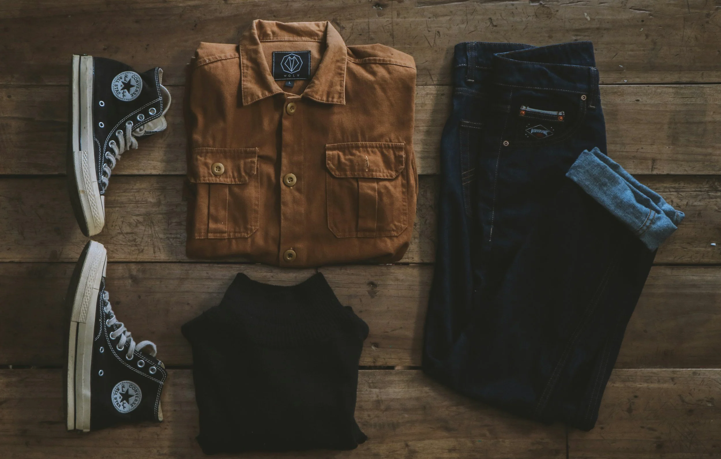 A pair of black high-top Converse sneakers on the left, a folded brown jacket in the center, with a black label that says 'WOLV', and a pair of dark blue jeans on the right, all laid out on a wooden surface with visible grain.