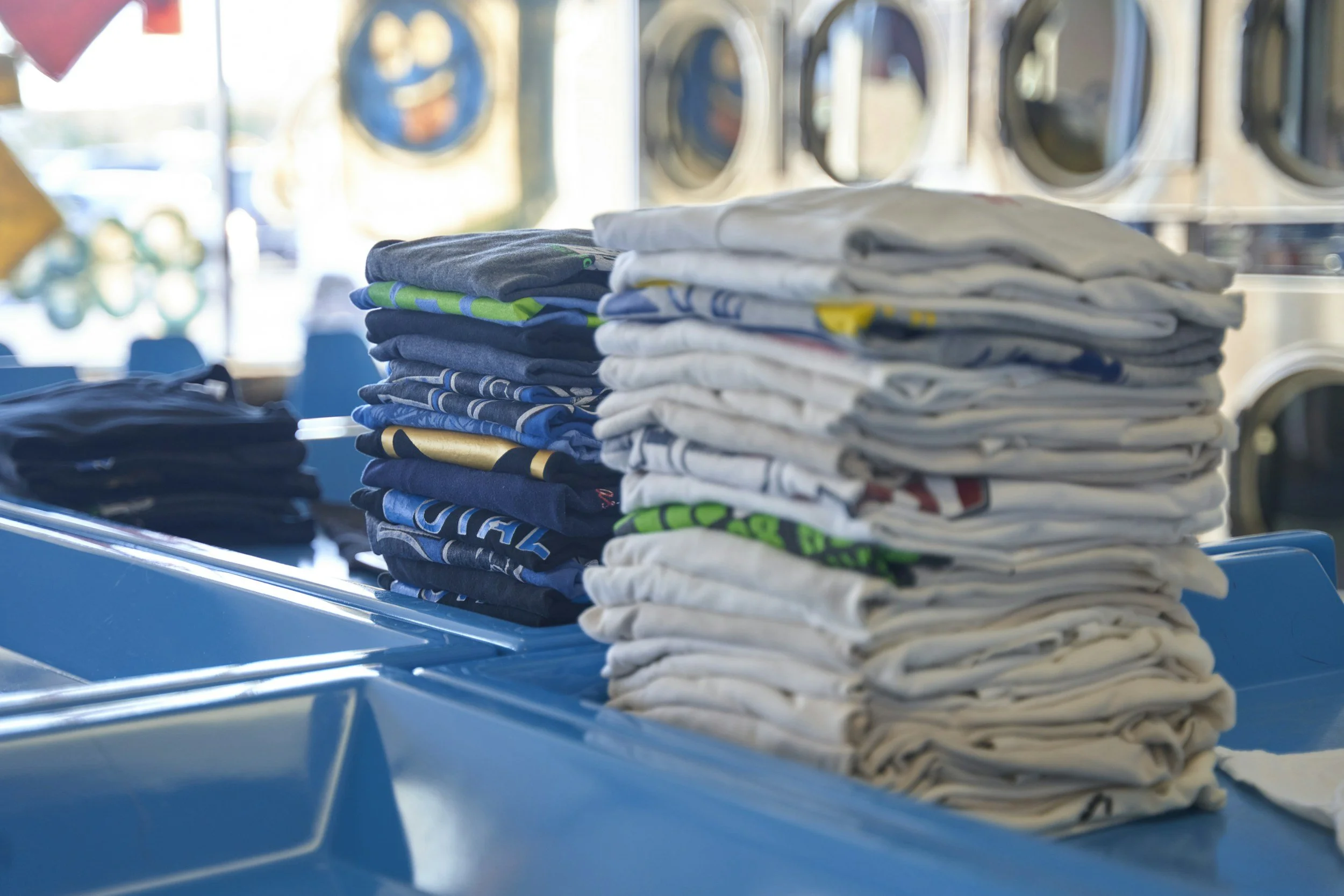 Stacks of folded T-shirts in various colors placed on a blue laundry cart in a laundromat with commercial washing machines in the background.