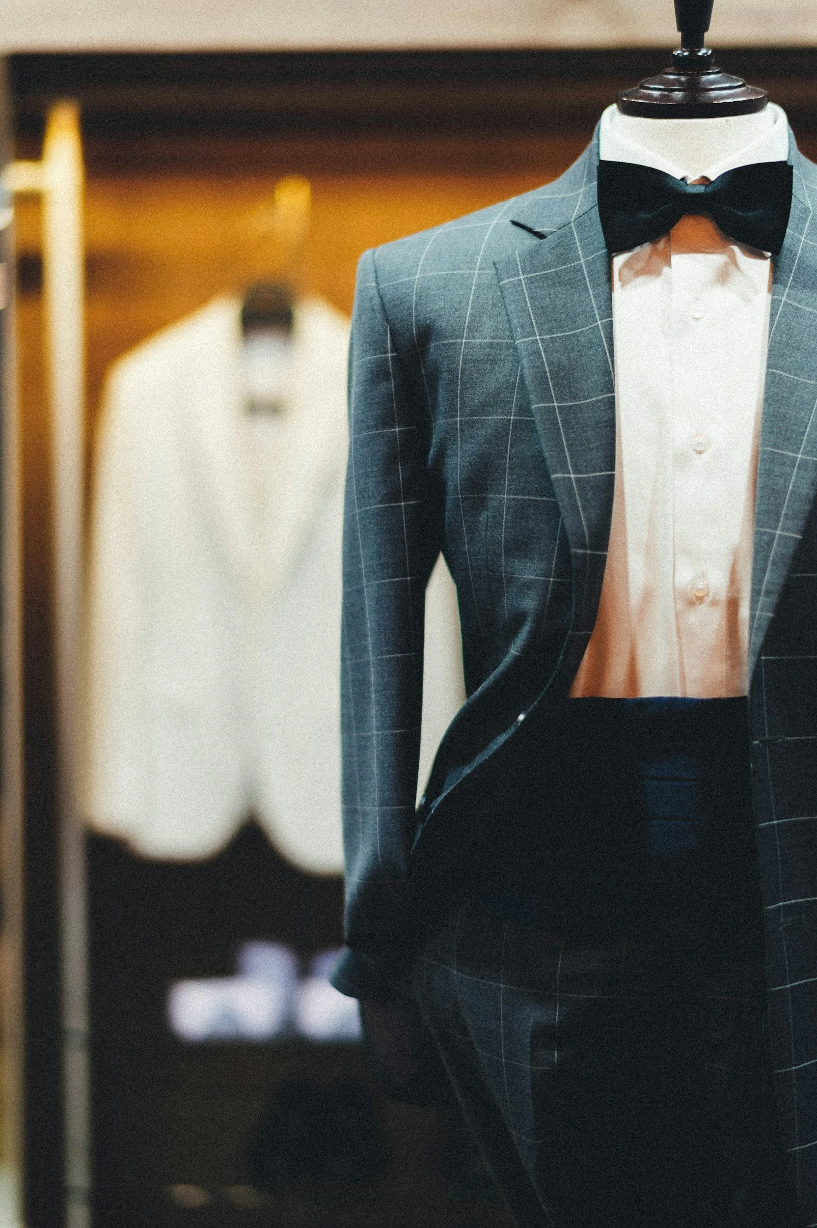 A mannequin dressed in a plaid suit with a white shirt and black bow tie, seen through a blurred mirror or glass reflection, with a rack of white shirts in the background.