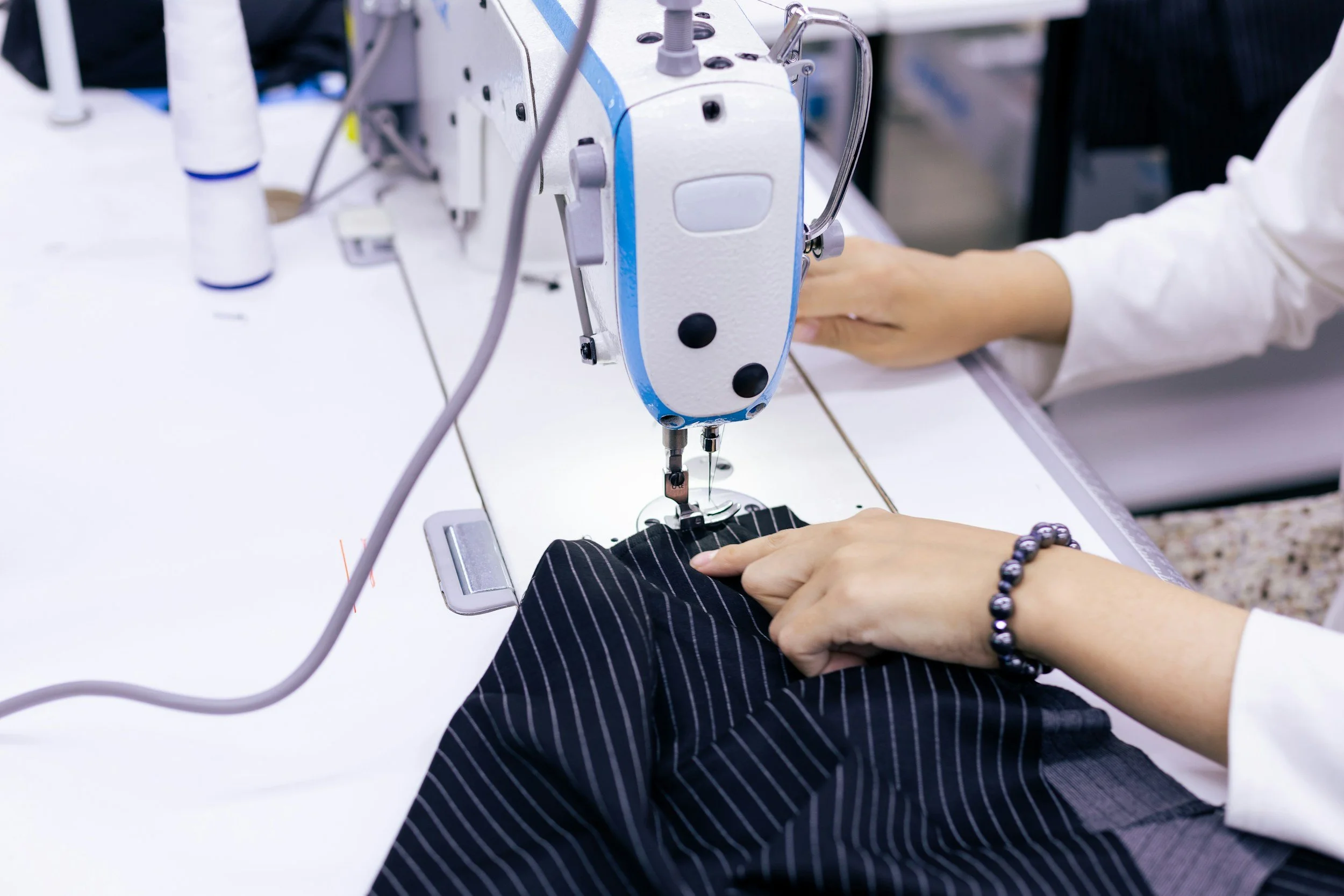 Close-up of a person sewing with a sewing machine, hands guiding black fabric with white pinstripes, wearing a bracelet and white sleeves.