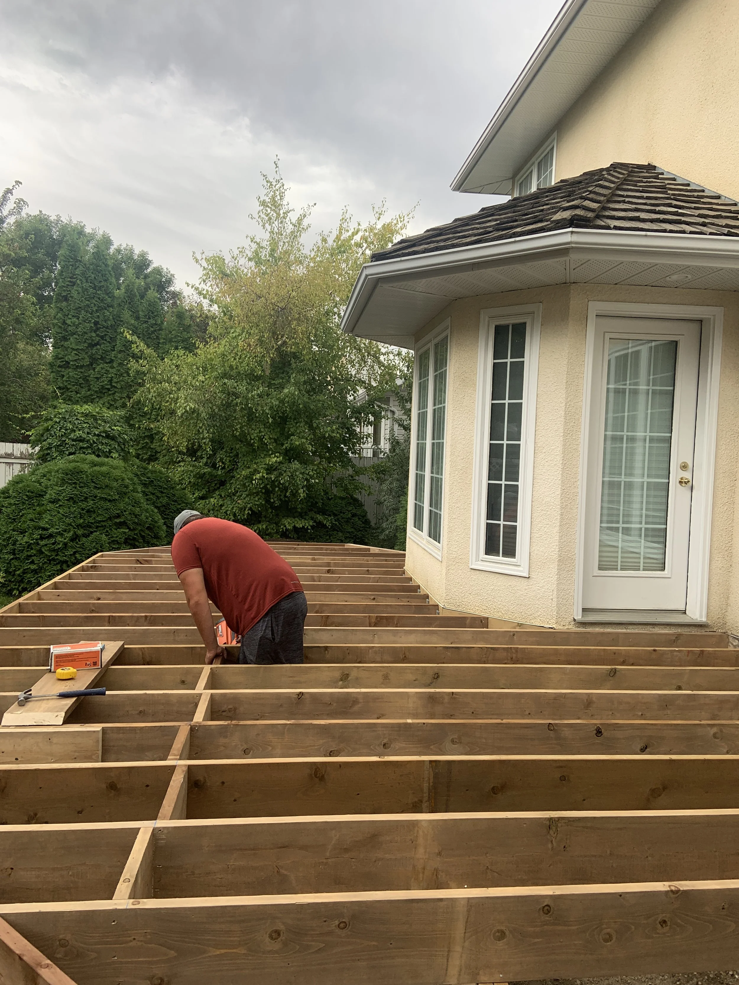 A person wearing a red shirt and shorts working on a wooden deck or platform beside a house, with tools and materials nearby, and trees in the background.