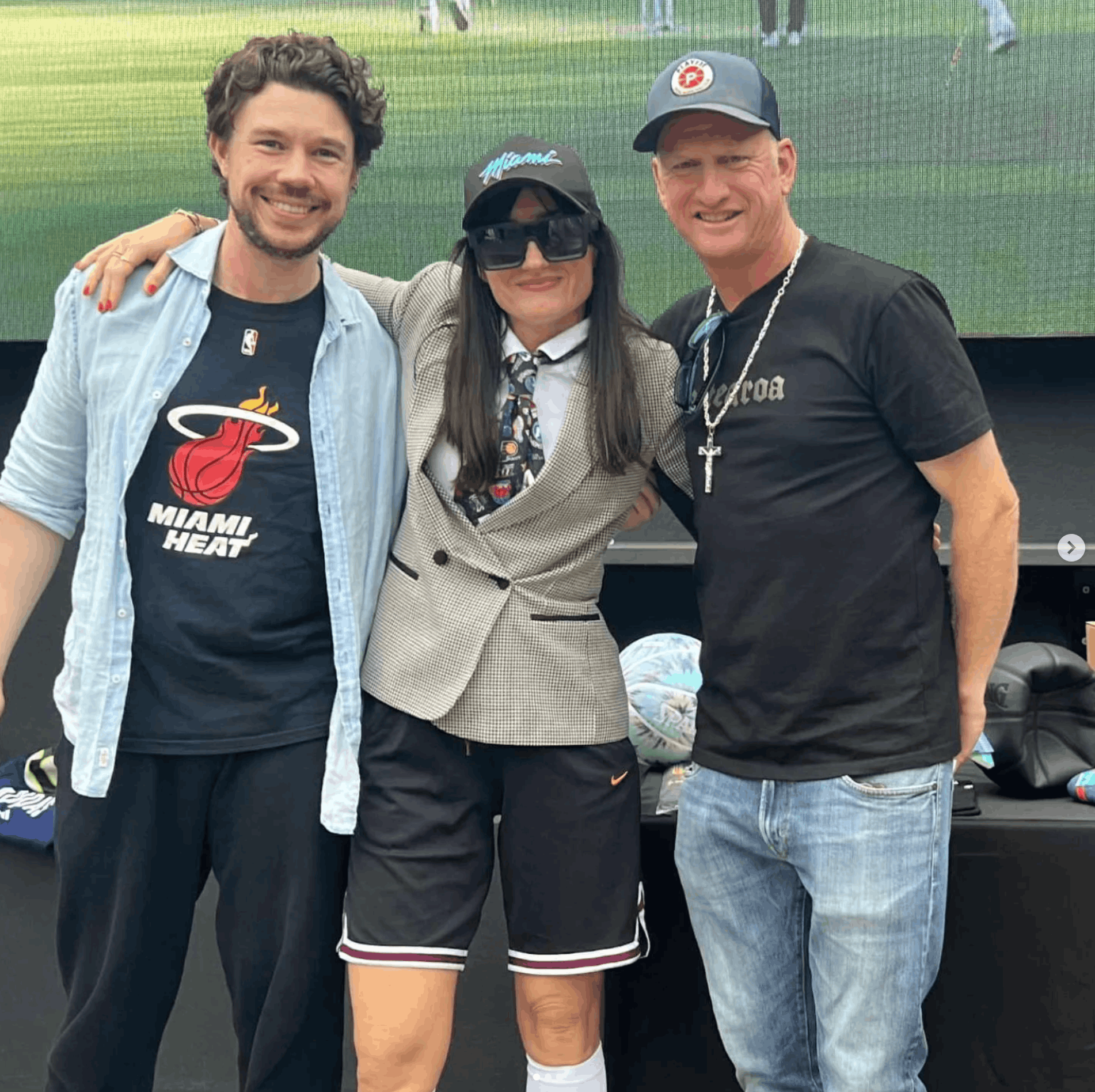 Three people standing close together outdoors. Two men and one woman are smiling at the camera. The woman is wearing dark sunglasses, a baseball cap, and a patterned blazer. The man on the right is wearing a black t-shirt and a chain necklace, and the man on the left is wearing a Miami Heat t-shirt and an unbuttoned denim shirt. The background shows a grassy area and people in the distance.
