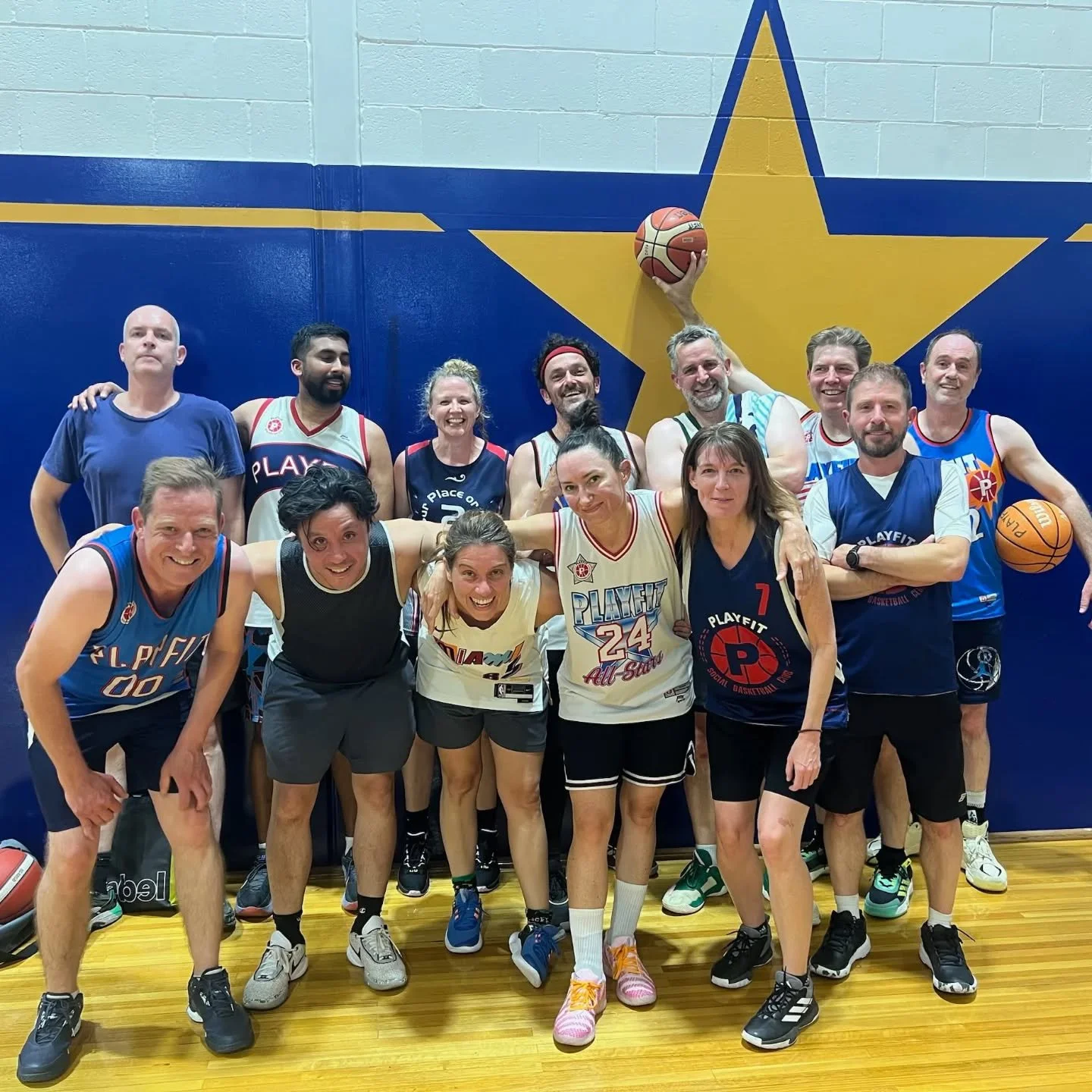 Group of people posing for a photo in an indoor basketball court with a large yellow star painted on the wall behind them. Some individuals are holding basketballs.