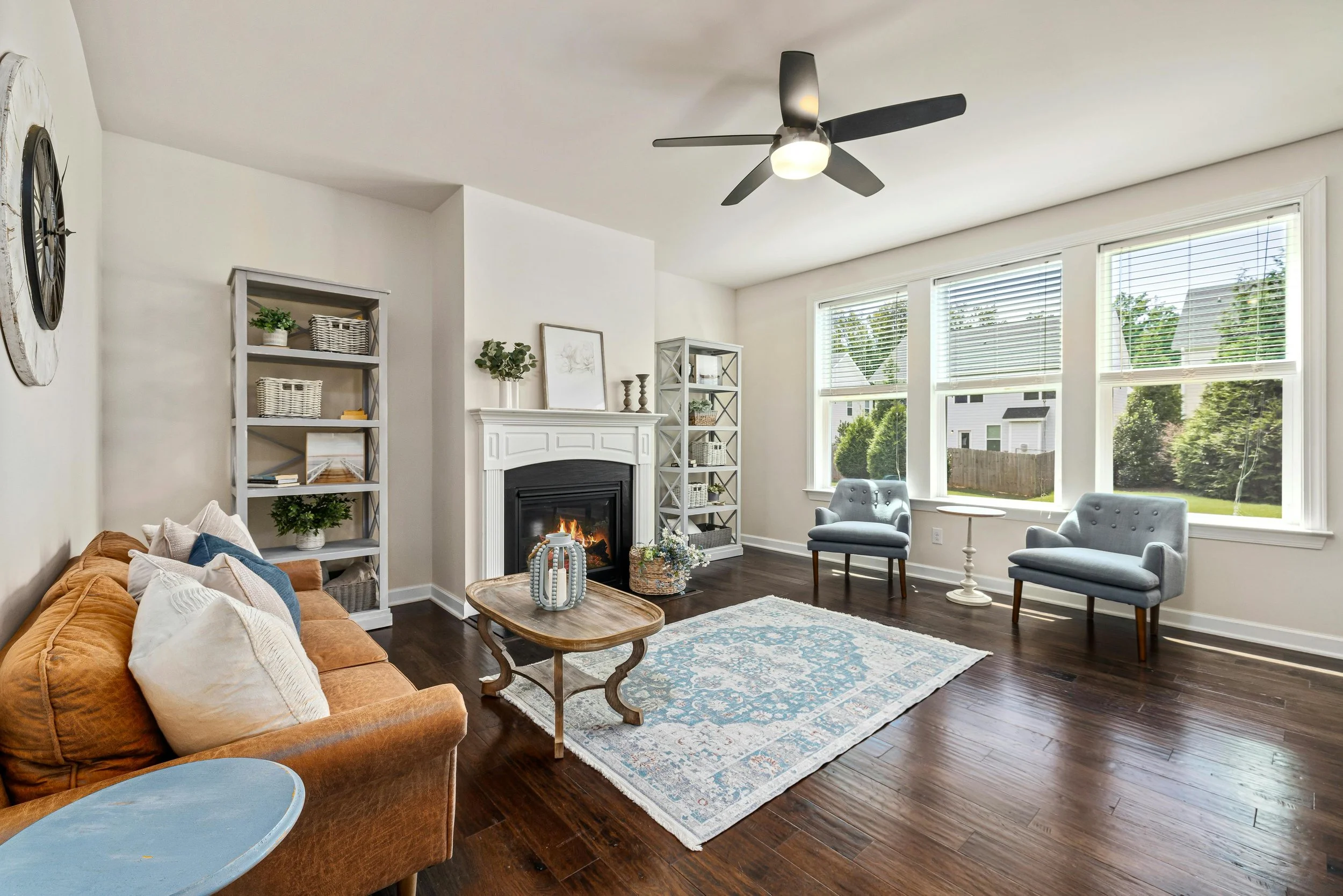 Bright living room with a fireplace, wooden floor, grey armchairs, a doll, bookshelf, large windows, and ceiling fan.