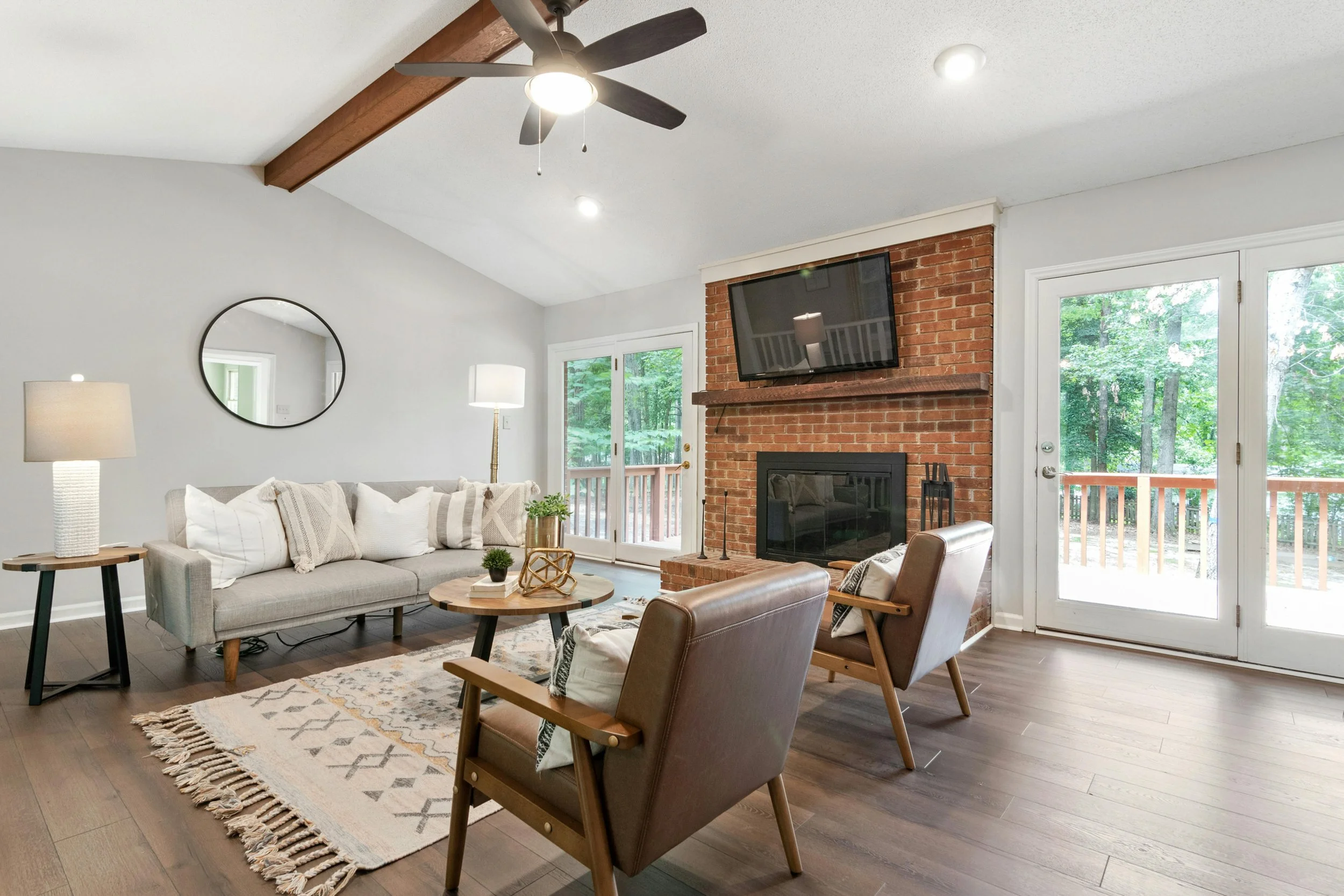 Living room with a gray sofa, two brown armchairs, a round wooden coffee table, a brick fireplace with a mounted TV, and large windows and doors showing a wooded outdoor area.