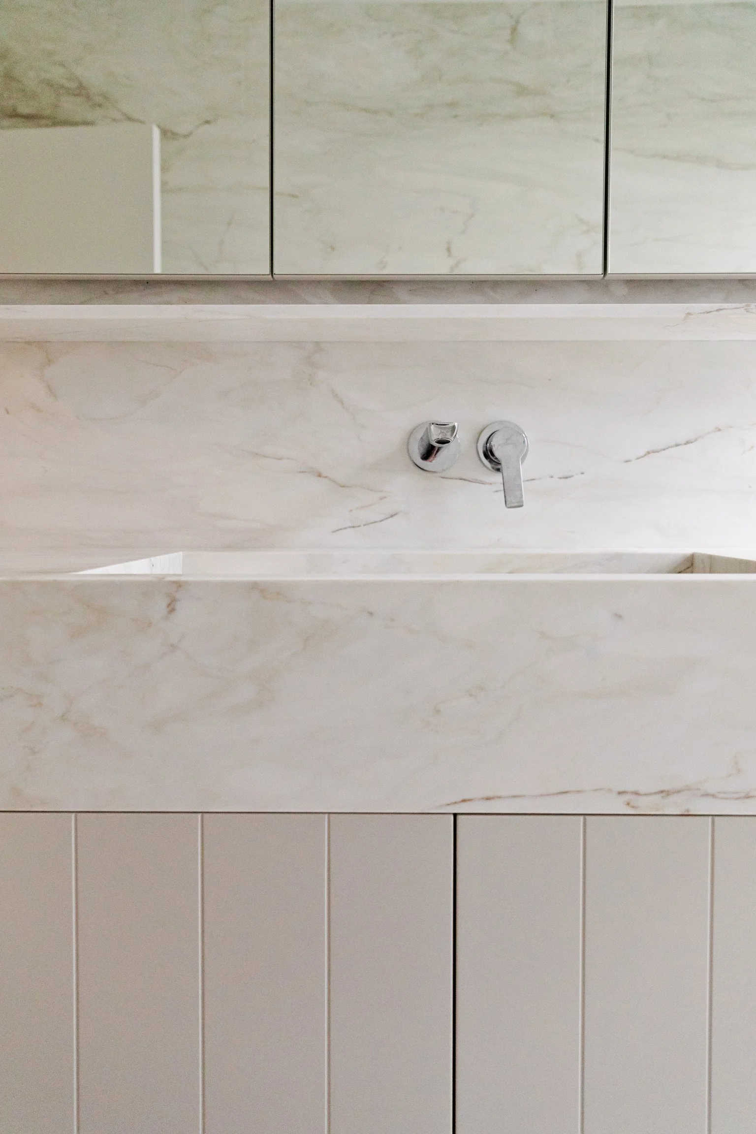 Bathroom wall with marble tiles, a double faucet, and a cabinet with mirror doors.