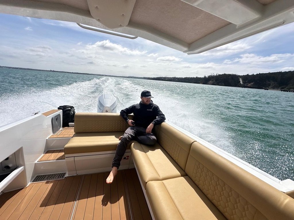 A man sitting on a tan cushioned bench on a boat, with water and distant land in the background.