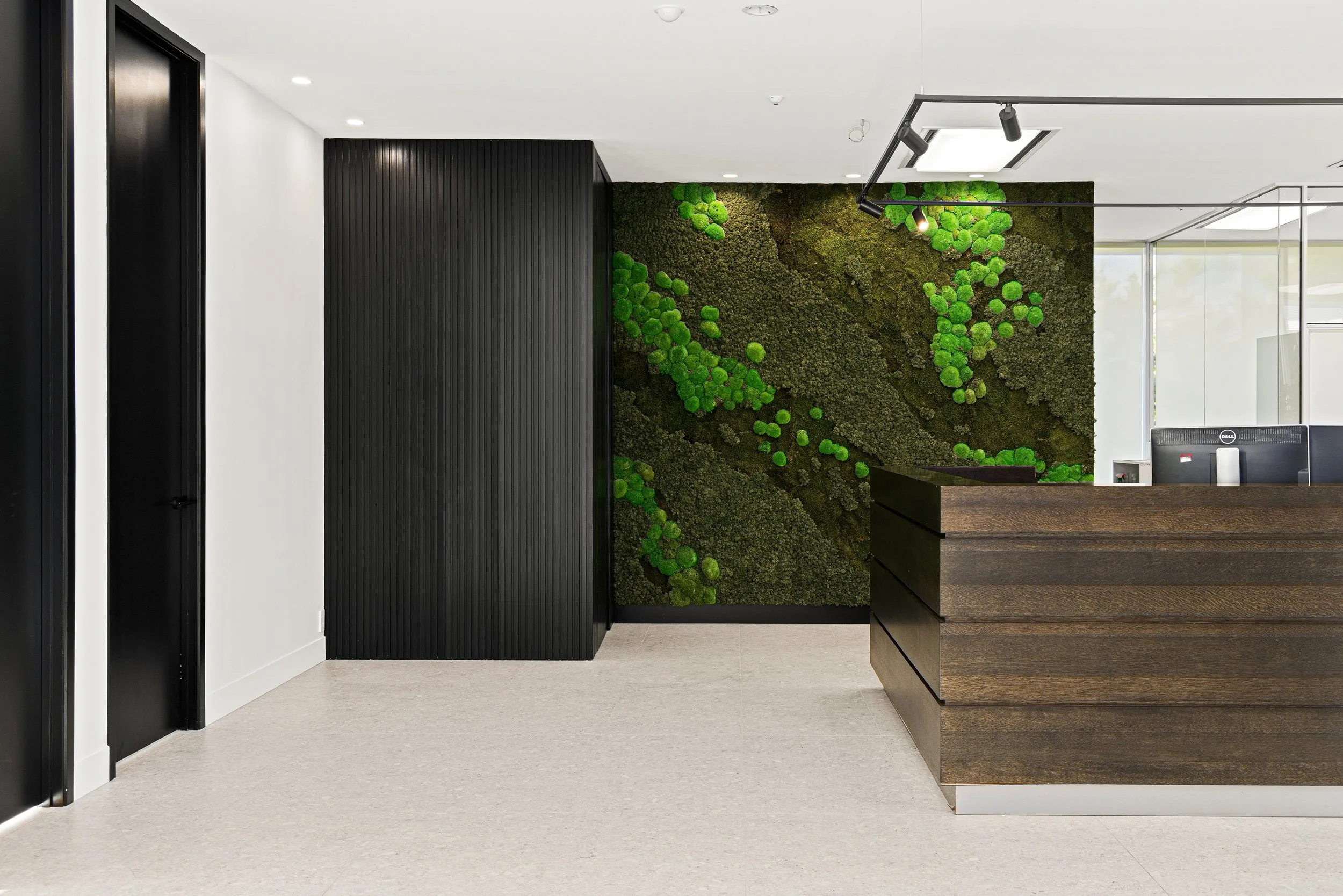 Modern office lobby with a green moss wall, a black vertical panel, a dark wooden reception desk, and black accents on doors and track lighting.