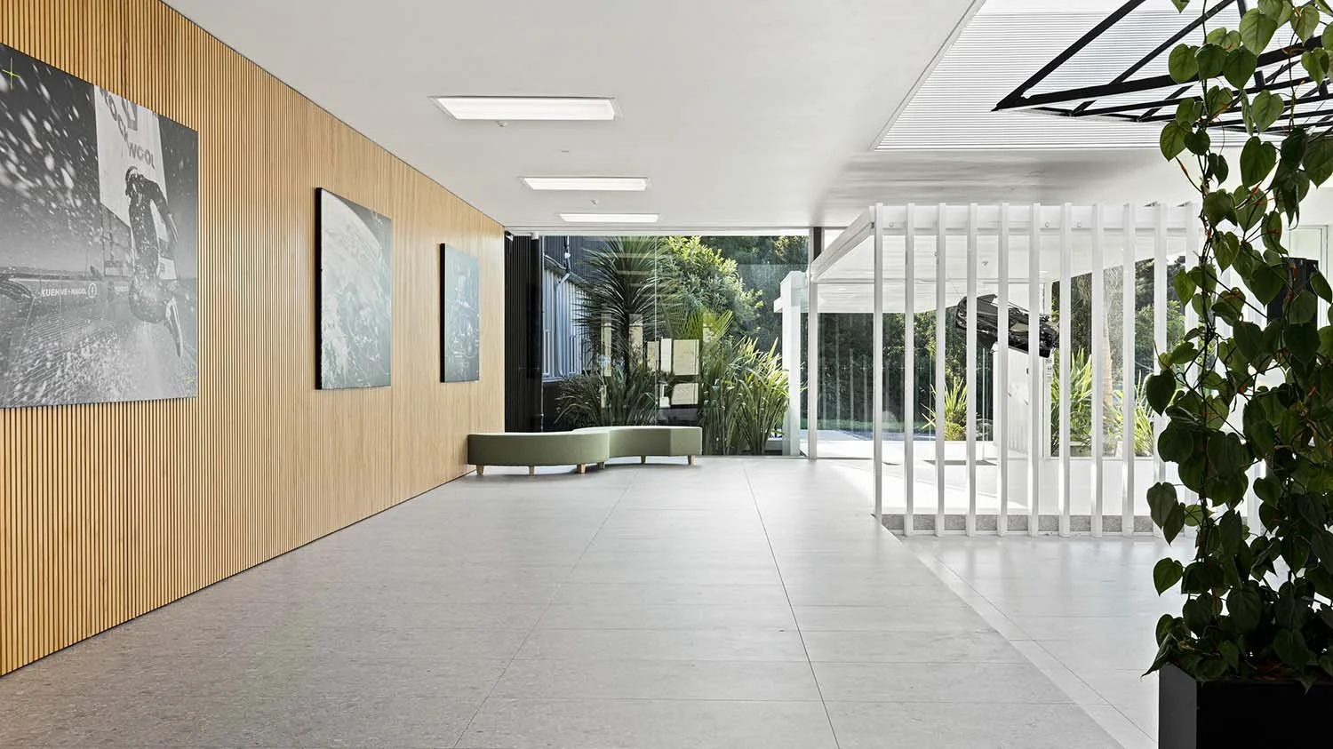 Interior space with wooden wall, modern art posters, large glass window, plants outside, green bench, and a white structure with bars and a car visible behind it.
