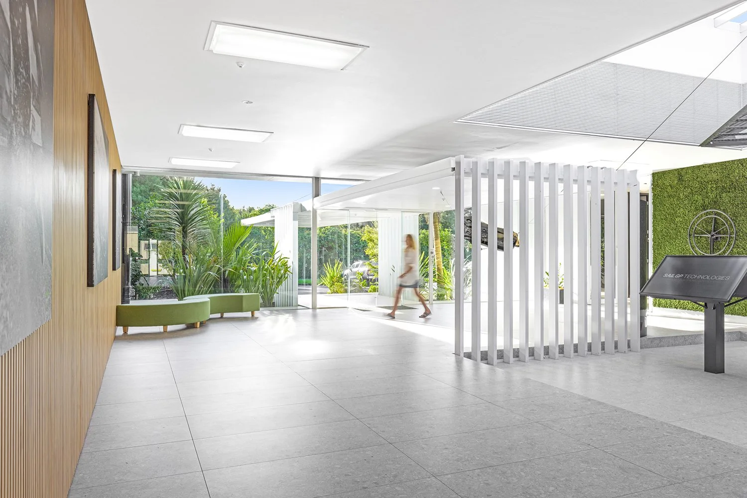 Modern lobby with large windows, green seating, indoor plants, and a digital info stand.