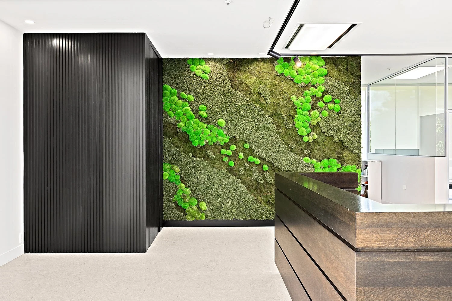 Modern interior with a moss and plant wall, black textured wall, wooden reception desk, glass walls, and bright lighting.
