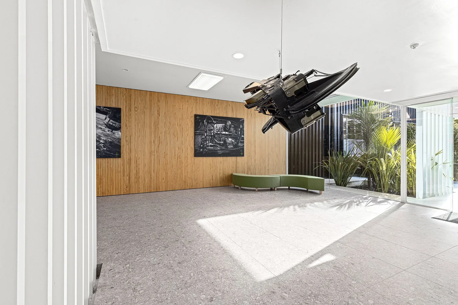 Modern lobby with wooden wall, green curved bench, large window with plants outside, and artwork on the wall; ceiling mounted mechanical object.