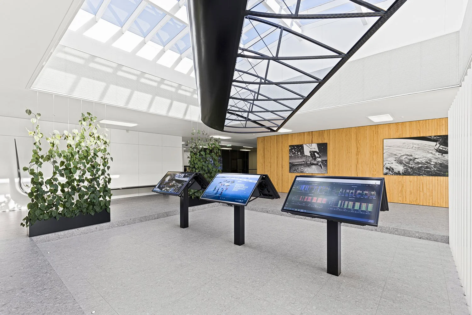 Modern interior space with three digital information screens on black stands, potted plants, wood paneled wall with black and white photographs, and a large curved skylight.