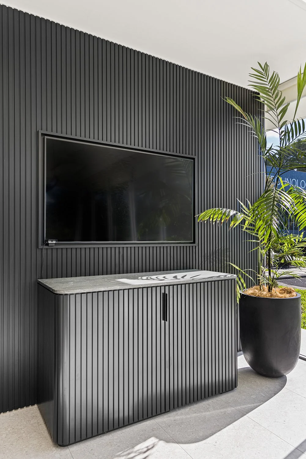 Modern black slatted accent wall with a large flat-screen TV and a black cabinet beneath it, next to a potted plant in a black planter, with sunlight casting shadows.