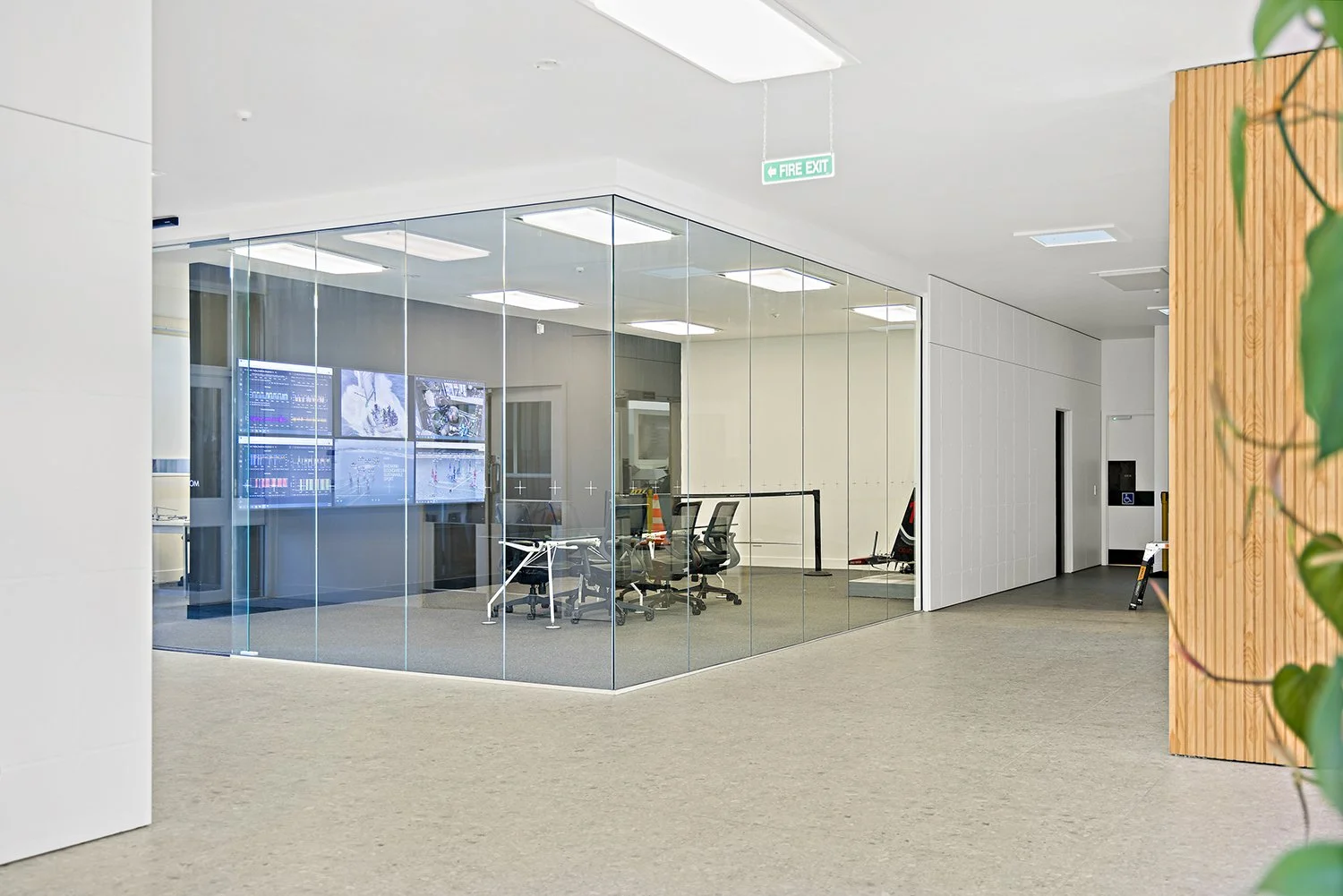 Modern office conference room with glass walls, multiple computer monitors displaying data, office chairs, and a white table, located inside a corporate building with a fire exit sign.