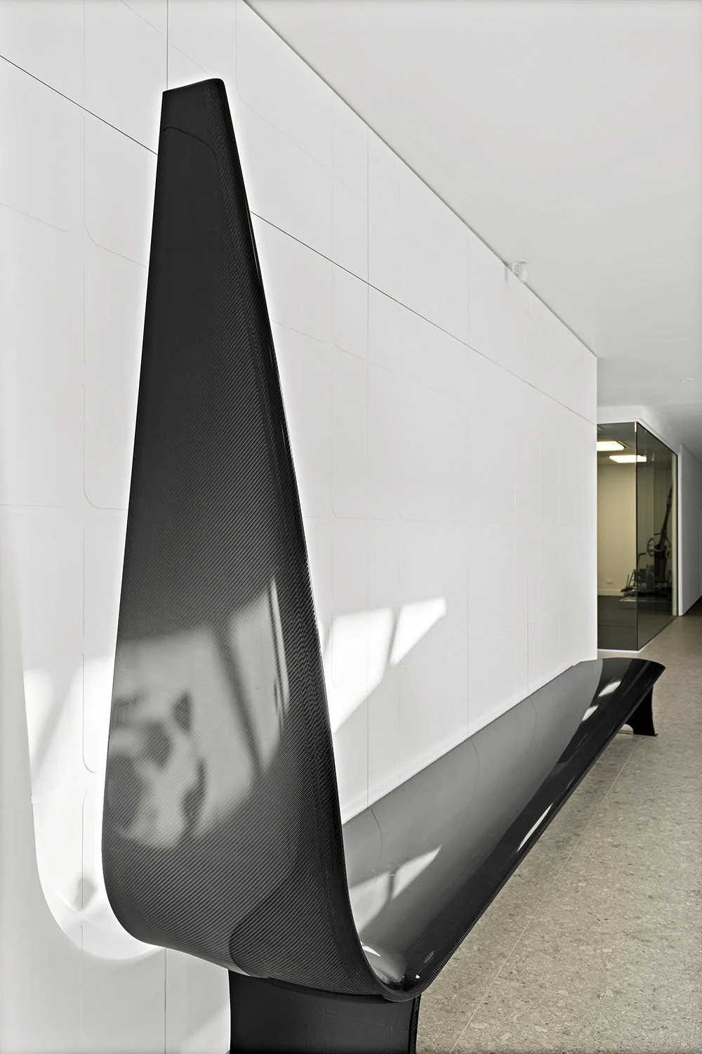 Modern black bench with a tall, curved backrest made of textured material, mounted against a white wall in a minimalistic indoor space.