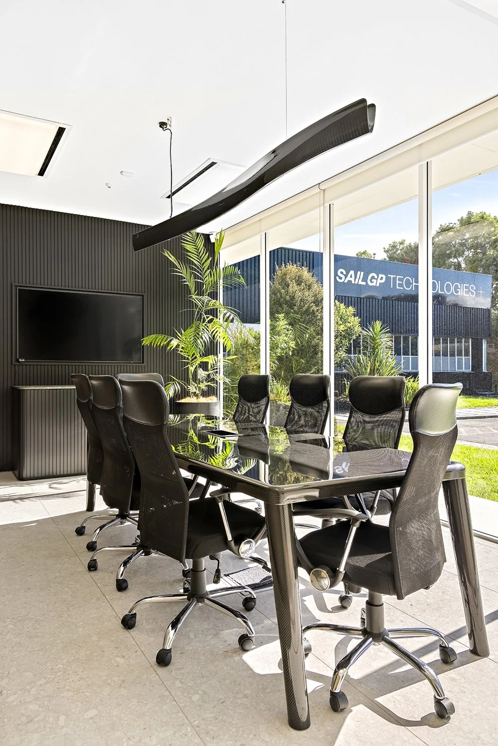 Modern conference room with a glossy black table, black office chairs, large windows, and a wall-mounted television, with outside view of a building labeled 'SAIL GP TECHNOLOGIES'.