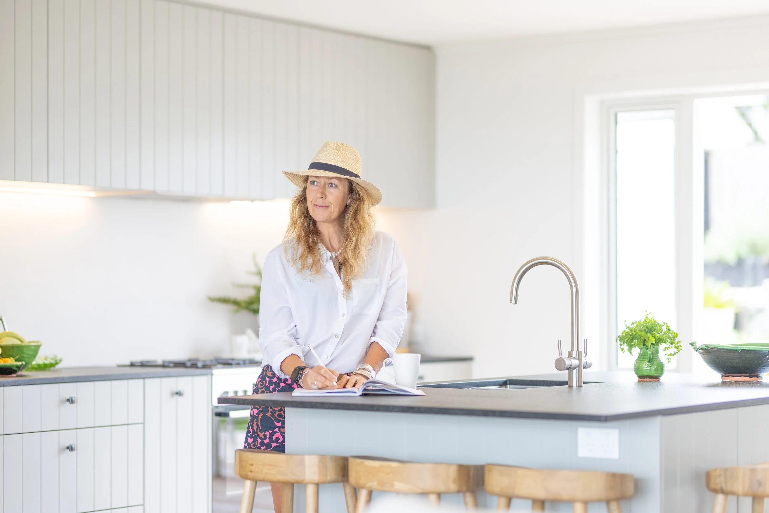 A woman with wavy blonde hair wearing a straw hat and a white shirt is standing in a bright, modern kitchen, looking to her left while writing in a notebook on a kitchen island.