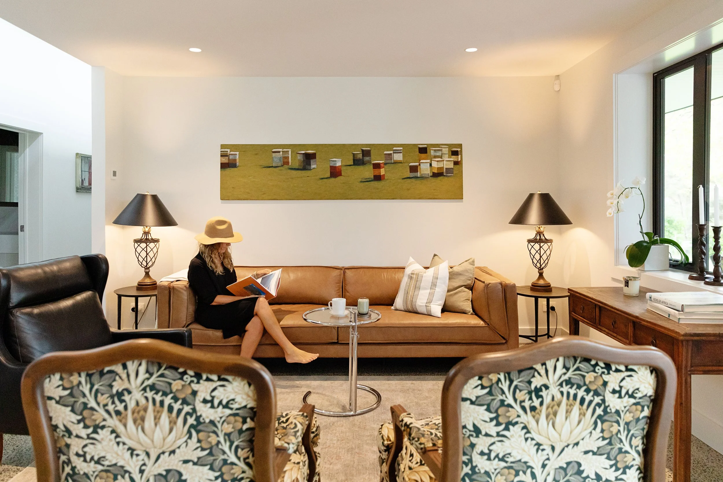 Living room with a woman sitting on a tan sofa reading a book, surrounded by patterned chairs, lamps, a coffee table with cups, and artwork on the walls, with large windows on the right letting in natural light.
