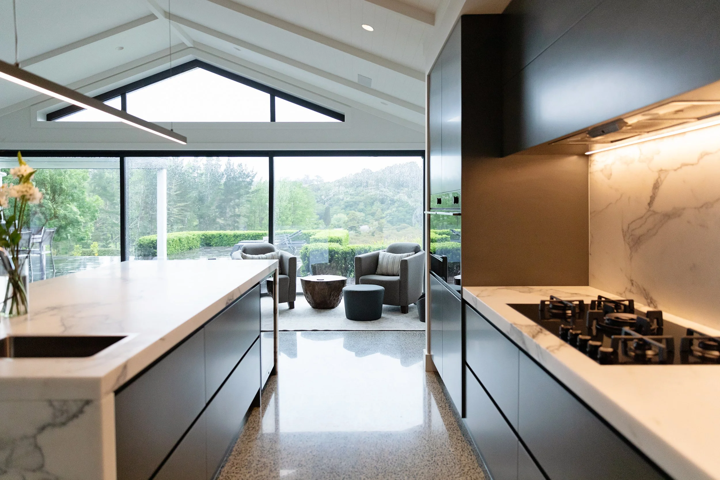 Modern kitchen with marble countertops and gas stove, view into a brightly lit living area with large windows, outdoor greenery, and seating area.