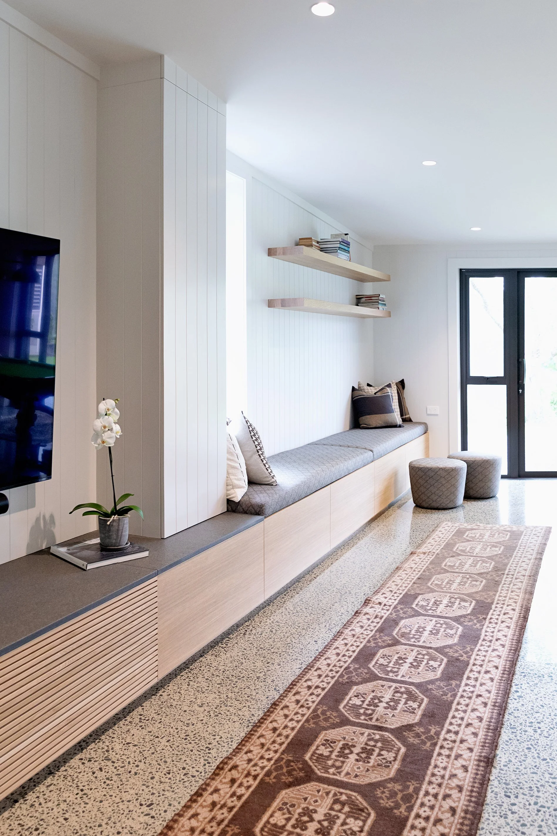 Modern living room with white paneled walls, floating wooden shelves with books, window seating with cushions, a patterned area rug, and a black door with glass panels.