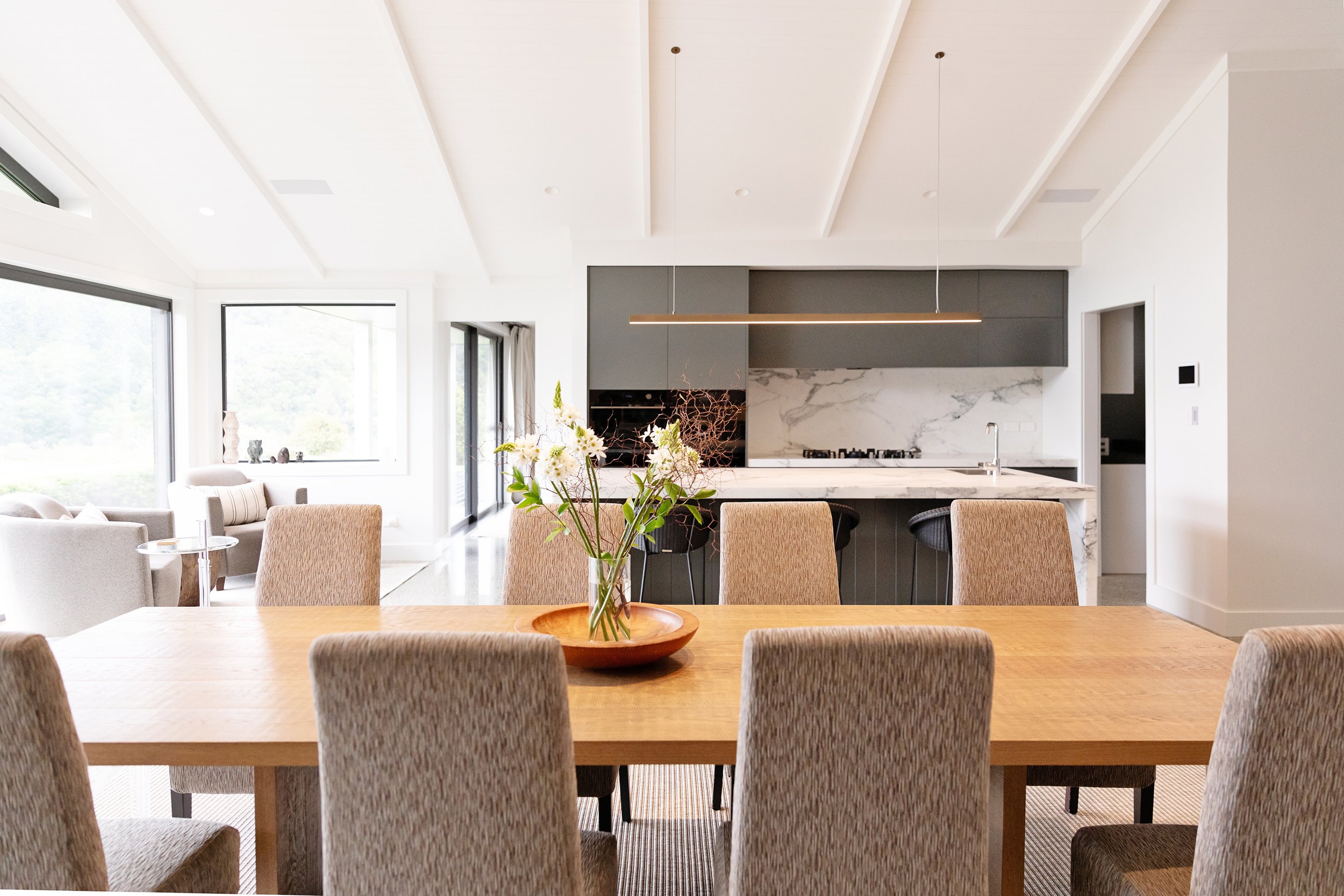 Modern open kitchen and dining area with a wooden table, beige upholstered chairs, a flower arrangement, large windows, and sleek gray cabinets.