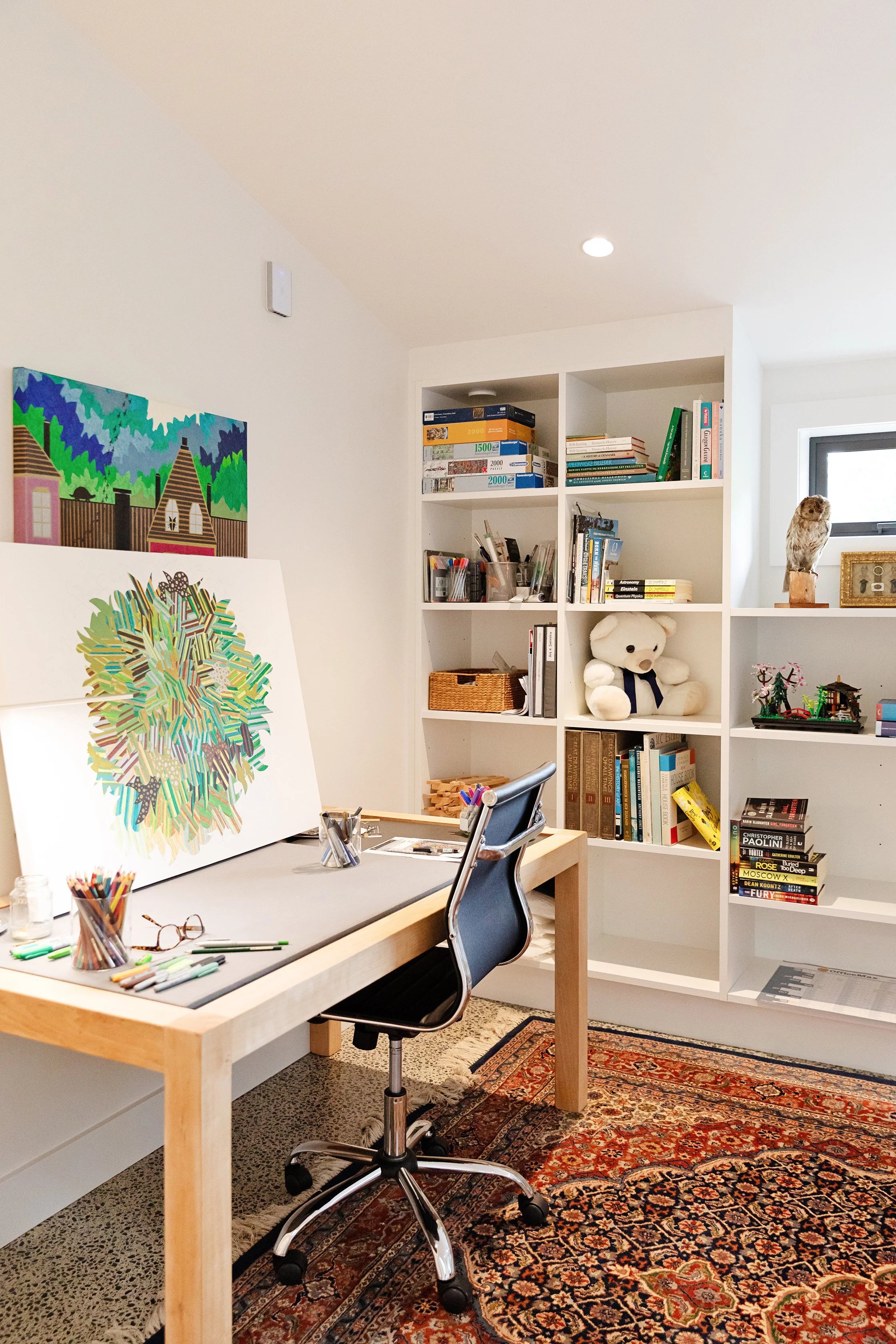 An art studio with a drawing desk, a computer chair, and art supplies including colorful markers. A large painting of an abstract tree is leaning against the wall. In the background, there is a white bookshelf with books, a teddy bear, and a small ow