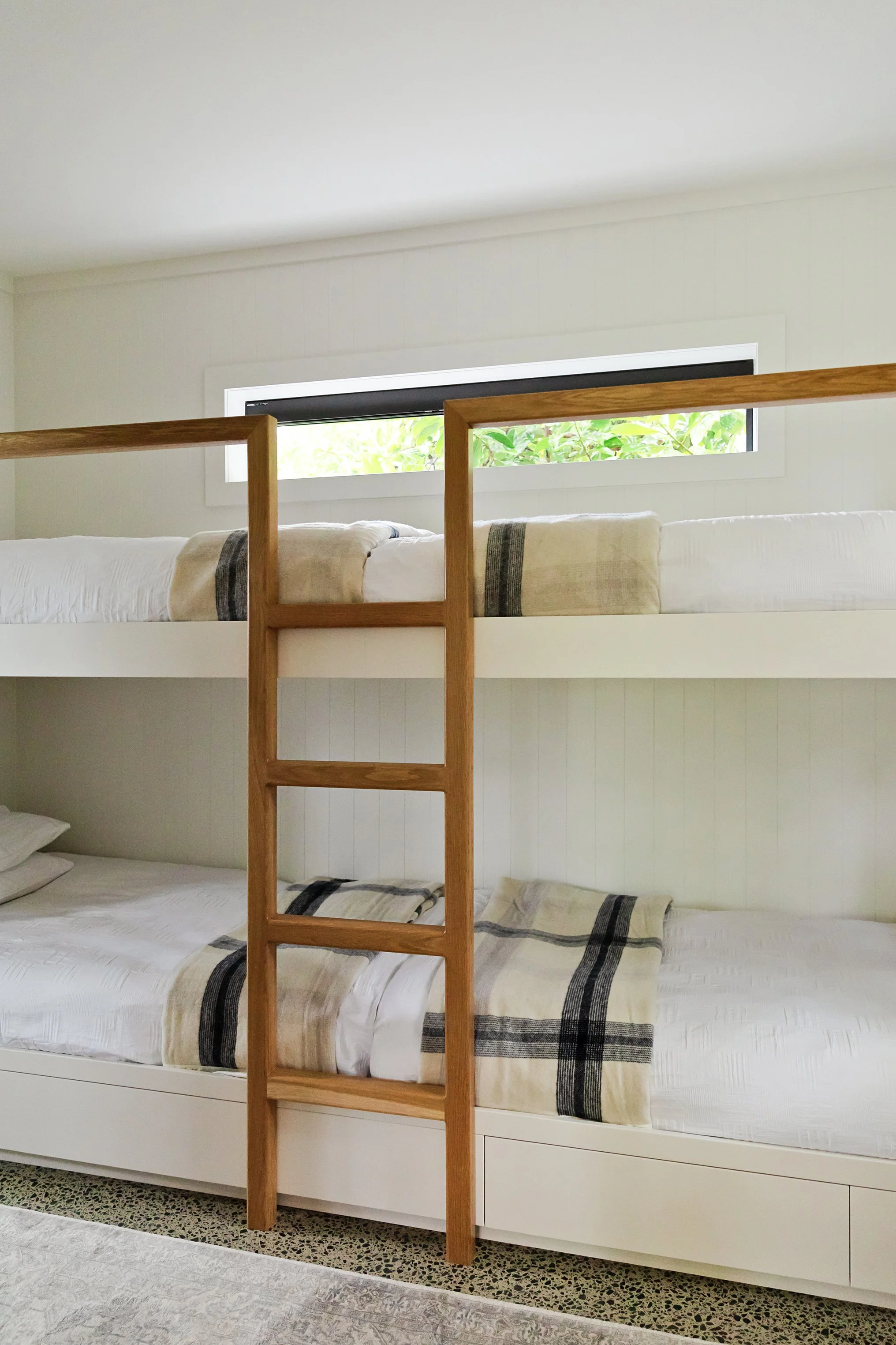 Bunk beds with white bedding and black-striped blankets, made with a ladder in the center, situated against a wall with a long, narrow window showing green foliage outside.