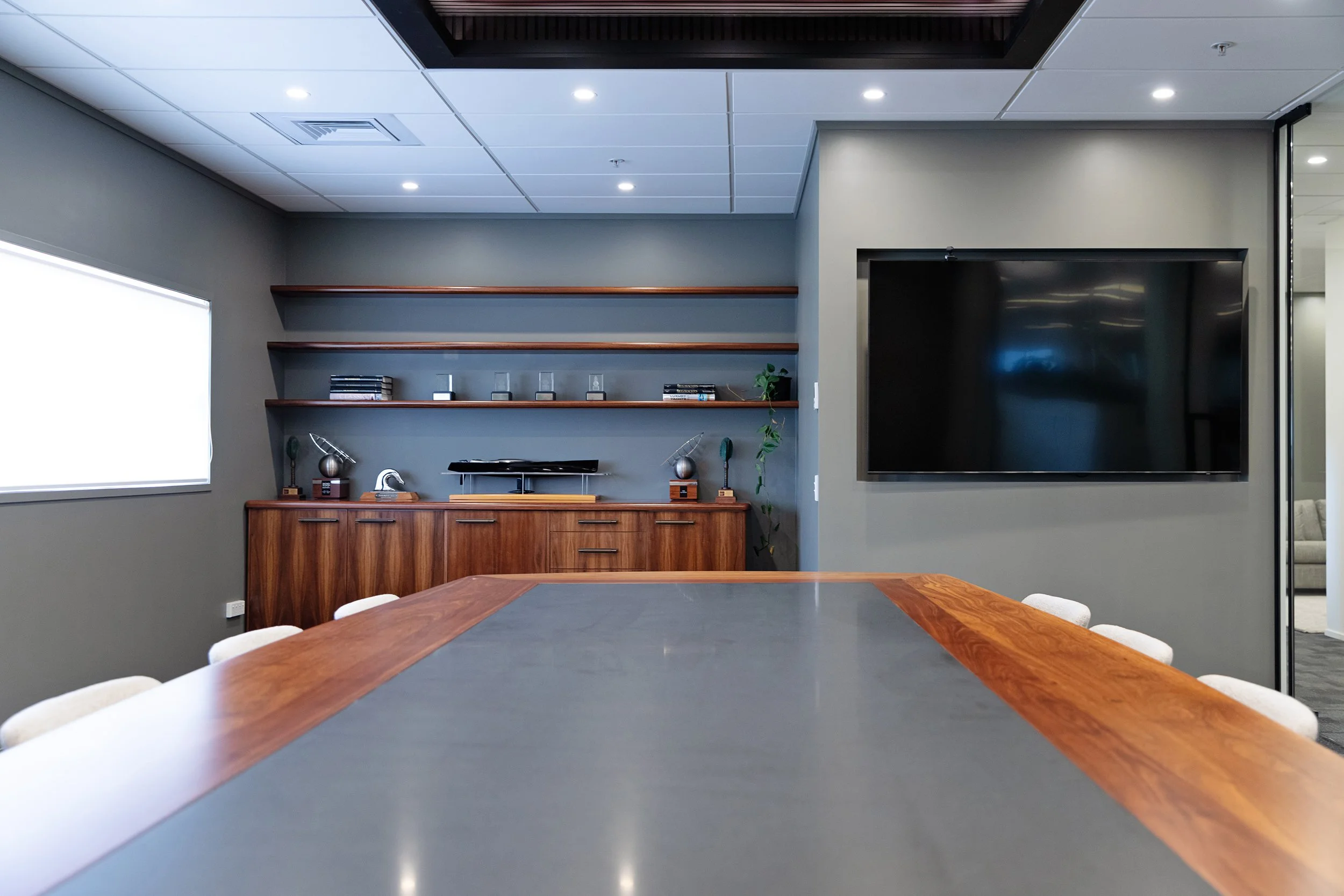 An office conference room with a large wooden table, beige chairs, grey walls, floating wooden shelves with decorative items, a large flat-screen TV, and a window letting in natural light.