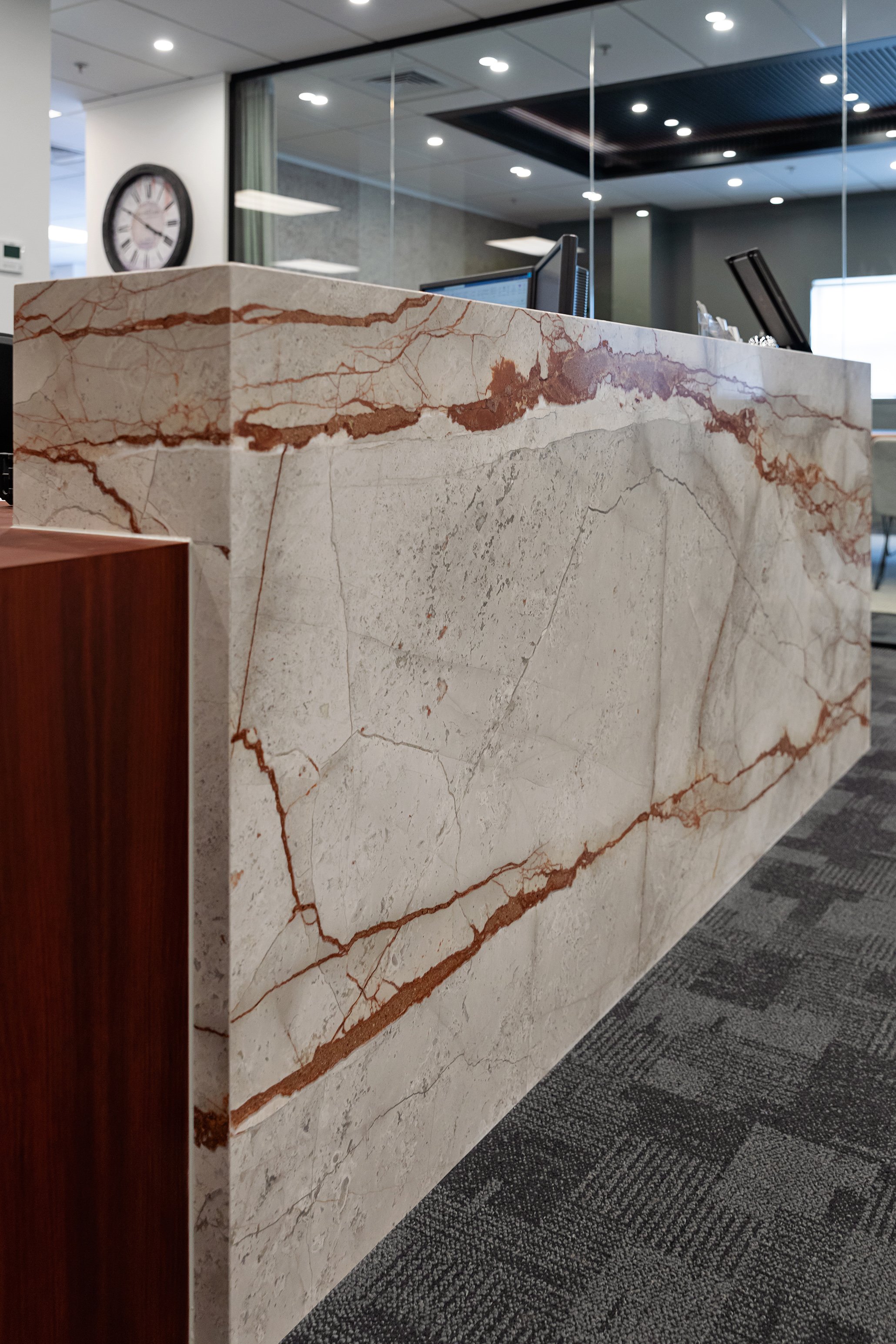 A marble reception desk with reddish-brown veining in an office lobby with glass partitions, a wall clock, and ceiling lights.