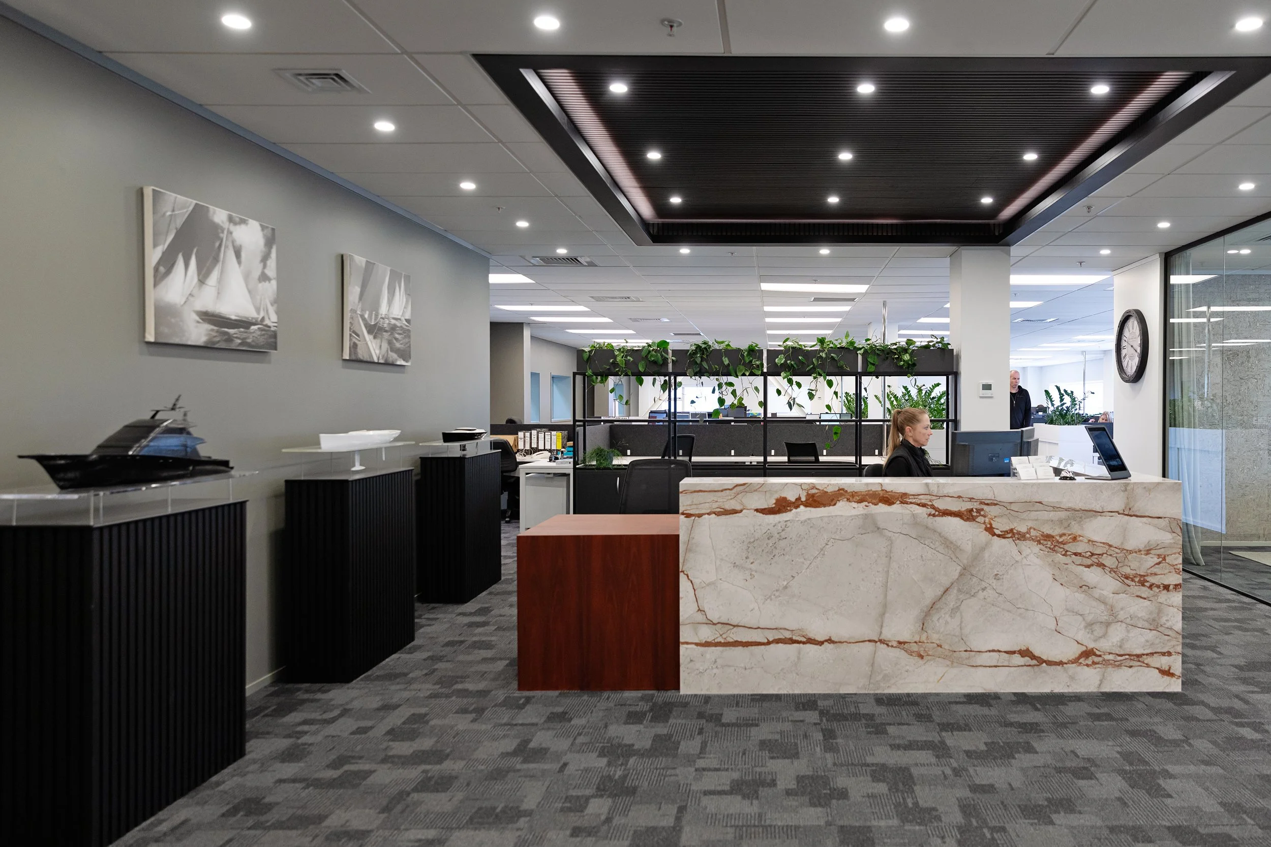 Modern office reception area with a marble counter, employees working at desks, potted plants, gray walls, artworks of sailboats, clock on glass wall, and a ceiling with recessed lighting.