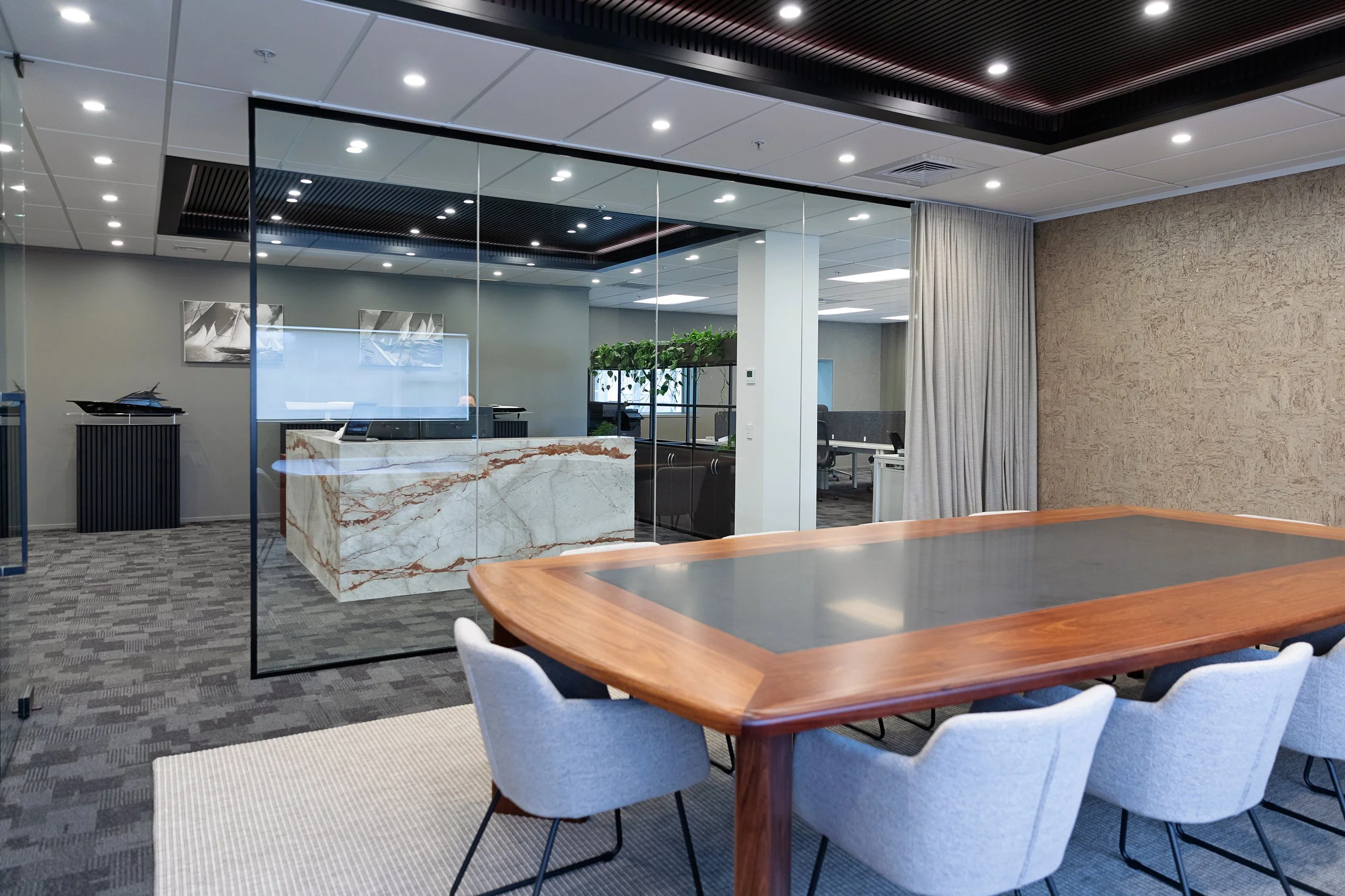 Modern office conference room with wooden table, light gray chairs, glass walls, ceiling lights, and a textured beige wall.