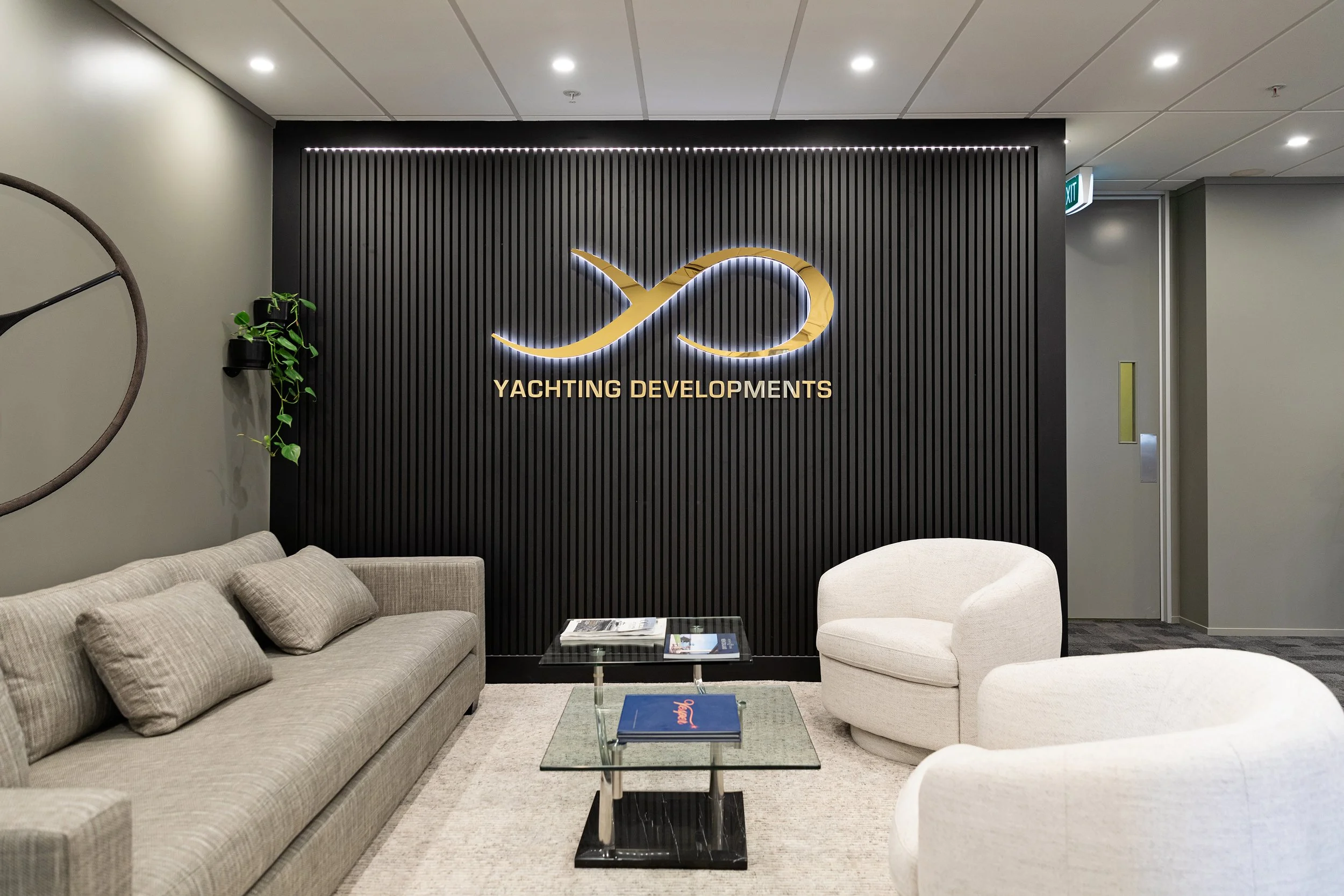 This is an indoor seating area with a gray couch and beige chairs around a glass coffee table. In the background, there is a black decorative wall panel with a gold logo and the words 'Yachting Developments' illuminated in gold. There are green plant