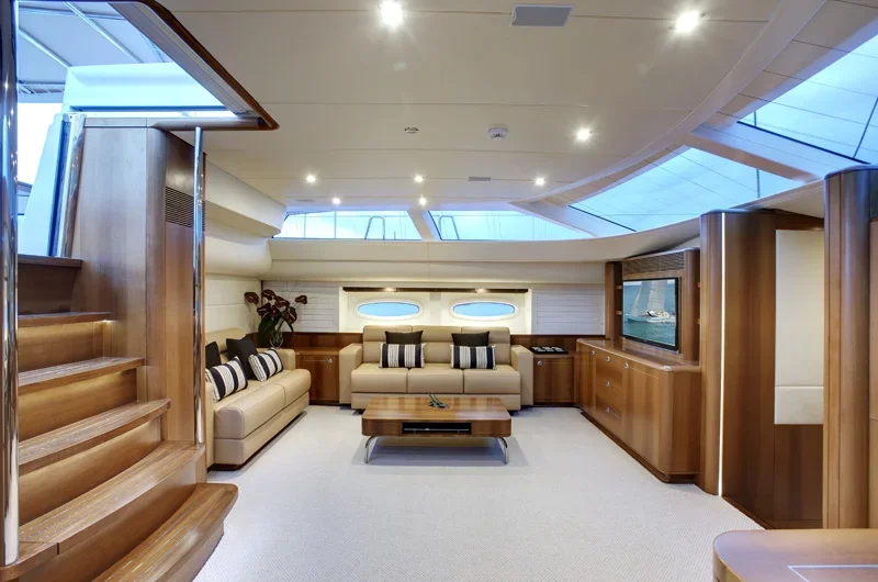 Interior of a luxurious yacht with beige sofas, a wooden coffee table, built-in cabinets, a flat-screen TV, and large windows, illuminated by ceiling lights.