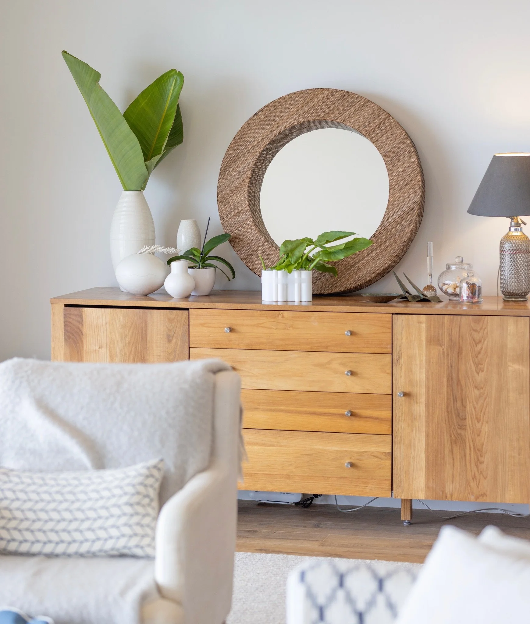 Wooden dresser with various decorated vases, plants, and bowls on top, a large round mirror hangs on the wall behind the dresser, a lamp on the right side, and part of a white sofa in the foreground.