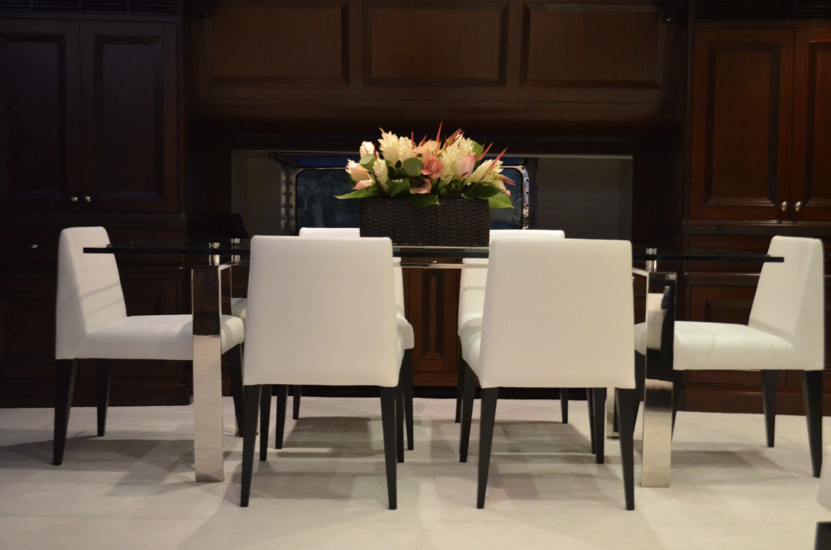A dining room with a round glass table, six white upholstered chairs, a rectangular floral centerpiece with pink and white flowers, and wooden cabinets in the background.