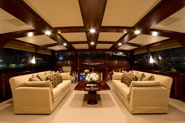 Interior of a yacht with two beige sofas, a wooden coffee table with a floral centerpiece, large windows, and recessed ceiling lights.