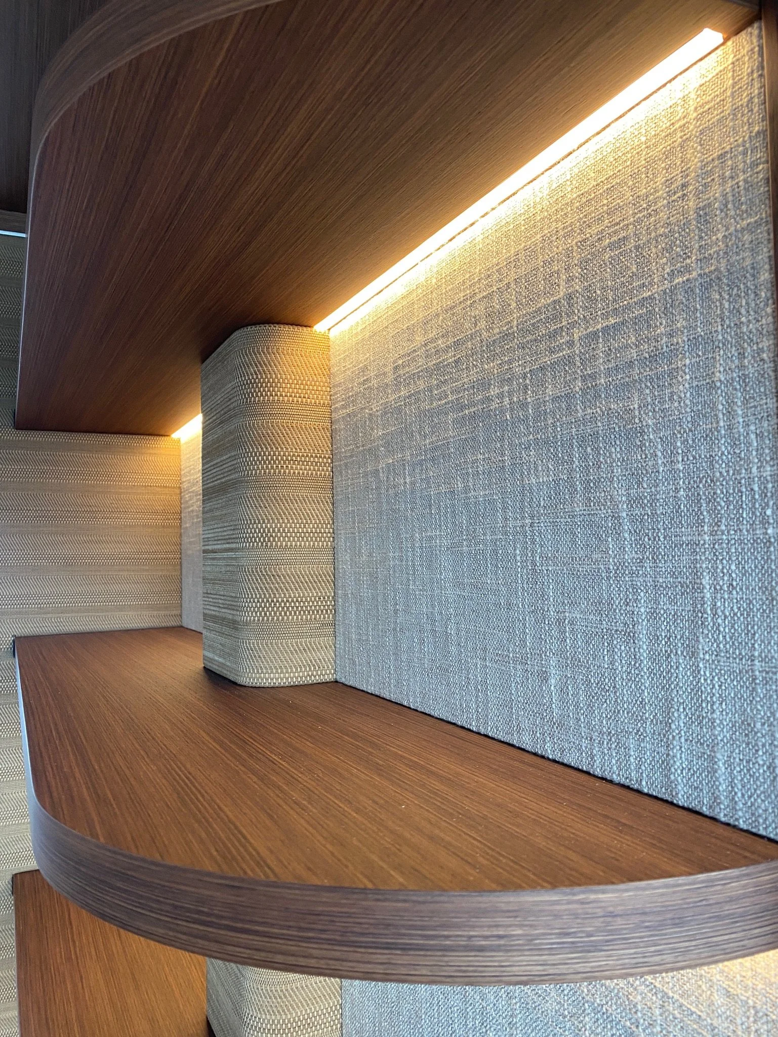 Close-up view of a modern curved wooden bookshelf with a fabric-covered wall behind it, featuring ambient lighting along the top edge.