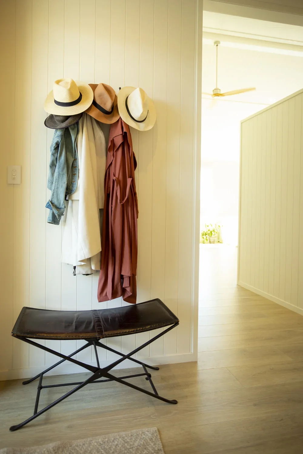 Wall with hooks holding hats, jackets, and dresses; foldable leather bench below; hallway with light-colored wood flooring and wall paneling.