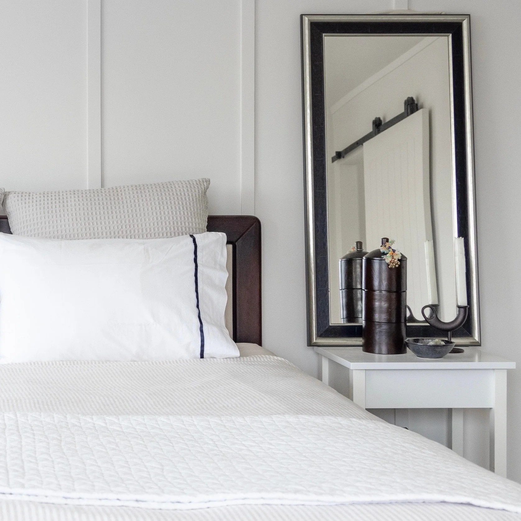 A neatly made bed with white bedding and a textured beige pillow, and a white nightstand next to it with decorative items, a large mirror on the wall, and a wall-mounted sliding door visible in the reflection.