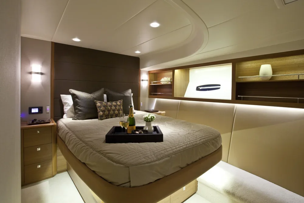 A modern bedroom with a large bed, dark headboard, and decorative pillows. There are built-in shelves, soft lighting behind a console, and a tray with drinks on the bed.