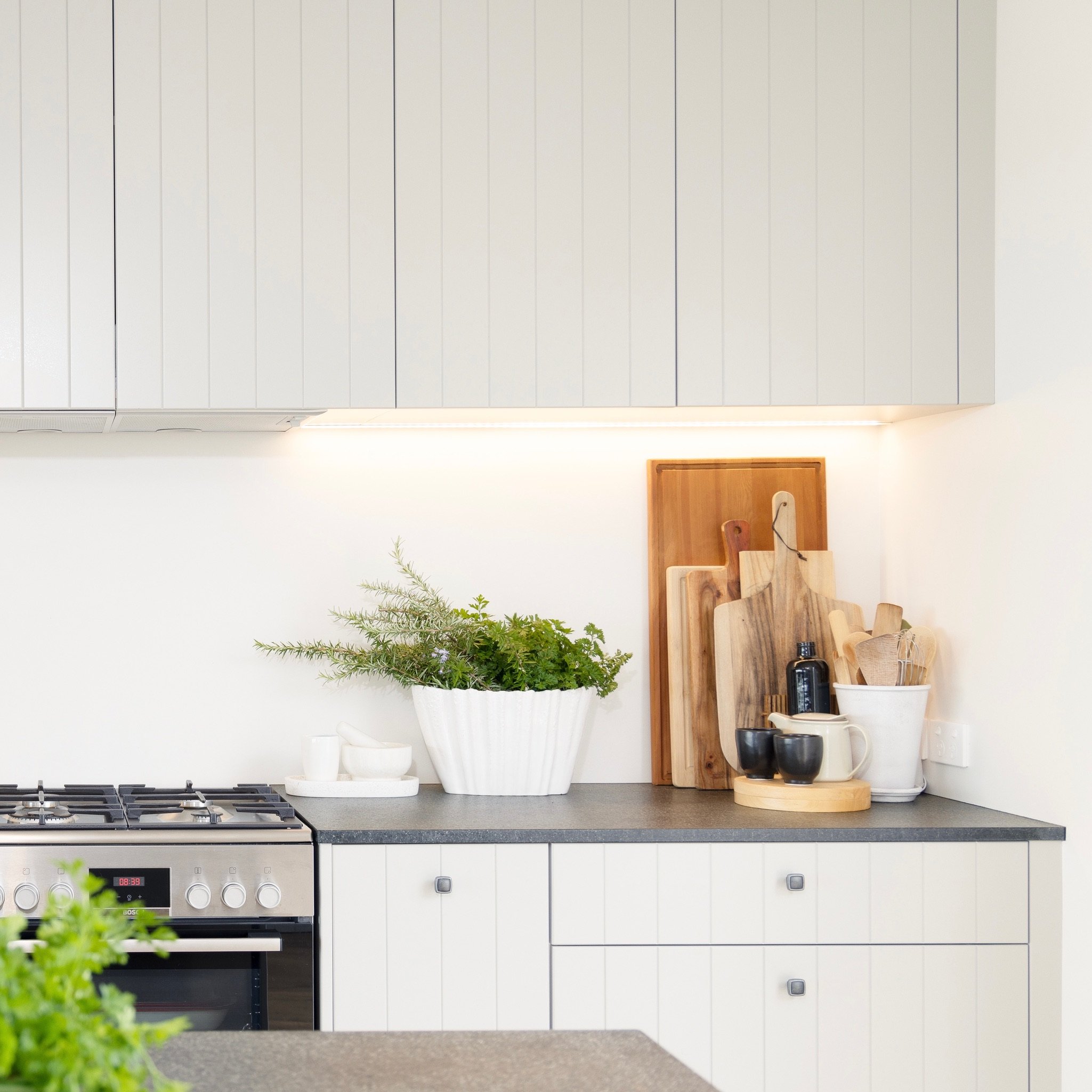 A kitchen countertop with a stove, white cabinets with vertical paneling, a wooden cutting board, various containers and a plant in a white pot.