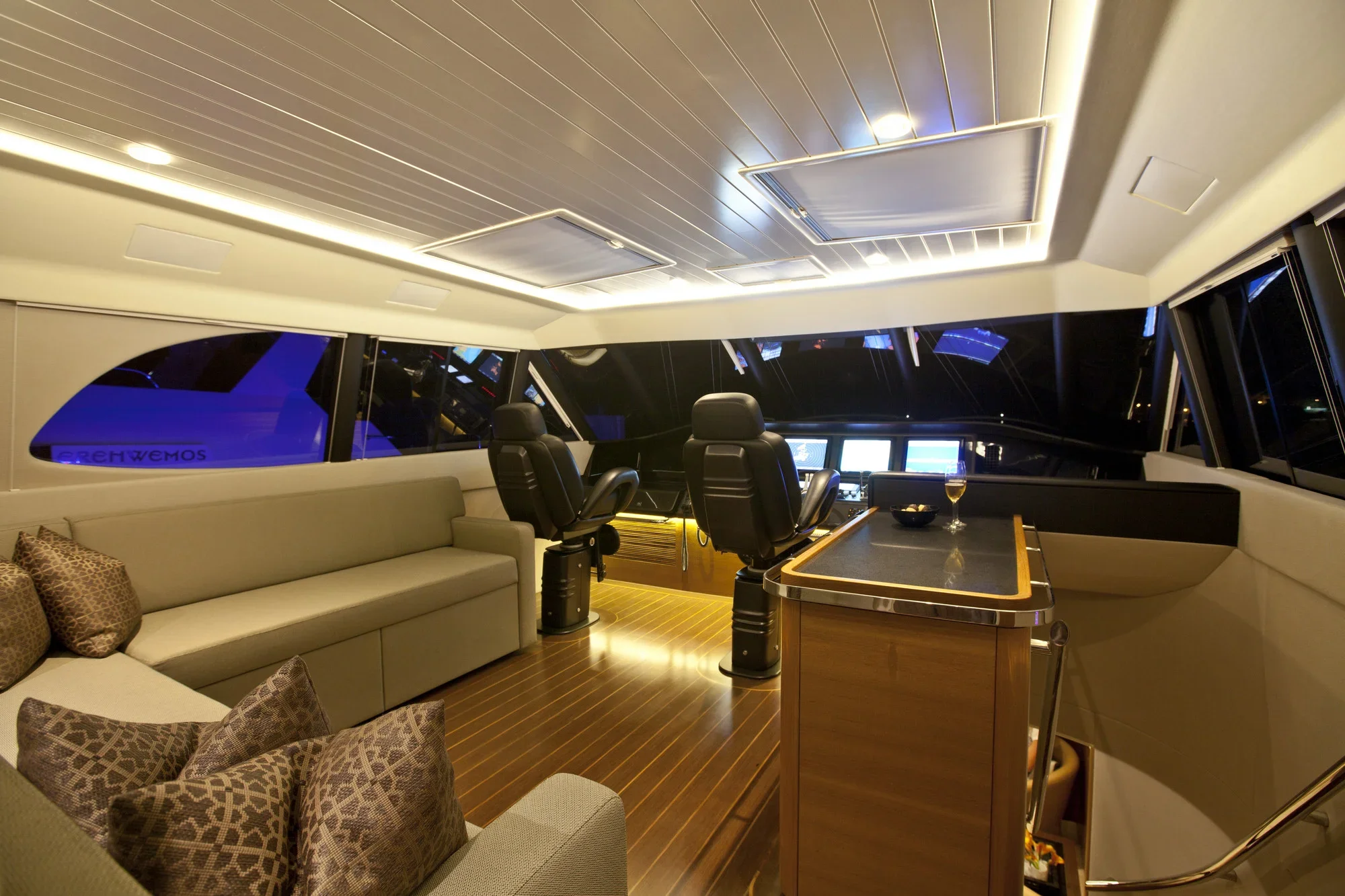 Interior shot of a yacht's control room with a beige sofa with patterned pillows on the left, multiple chairs facing electronics and steering controls, and large windows showing night outside.