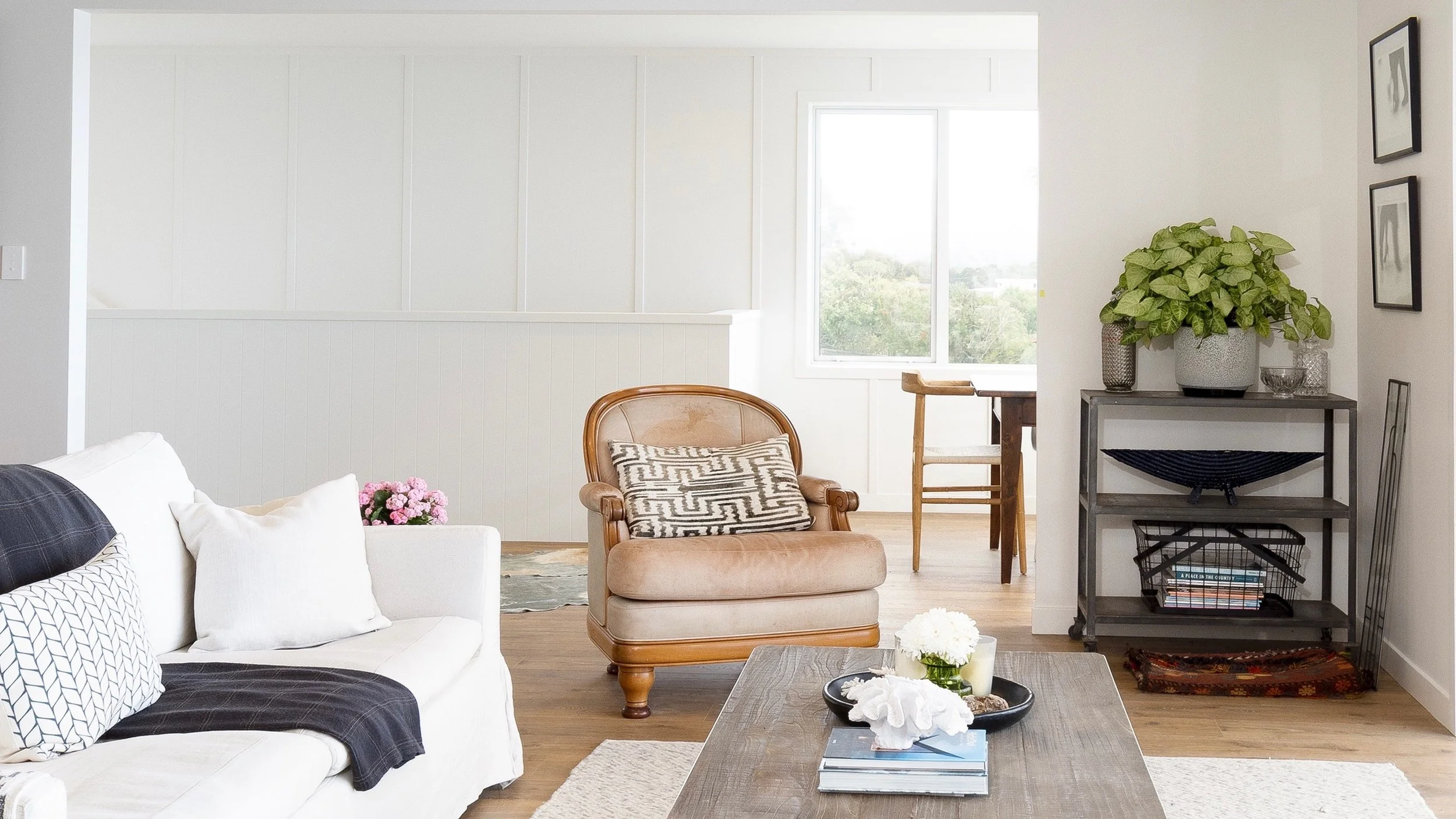 Interior of a modern living room featuring a white sofa with patterned cushions, a wooden armchair with a black and white patterned cushion, a coffee table with books and decorative items, a console table with potted plants and framed photos on the w