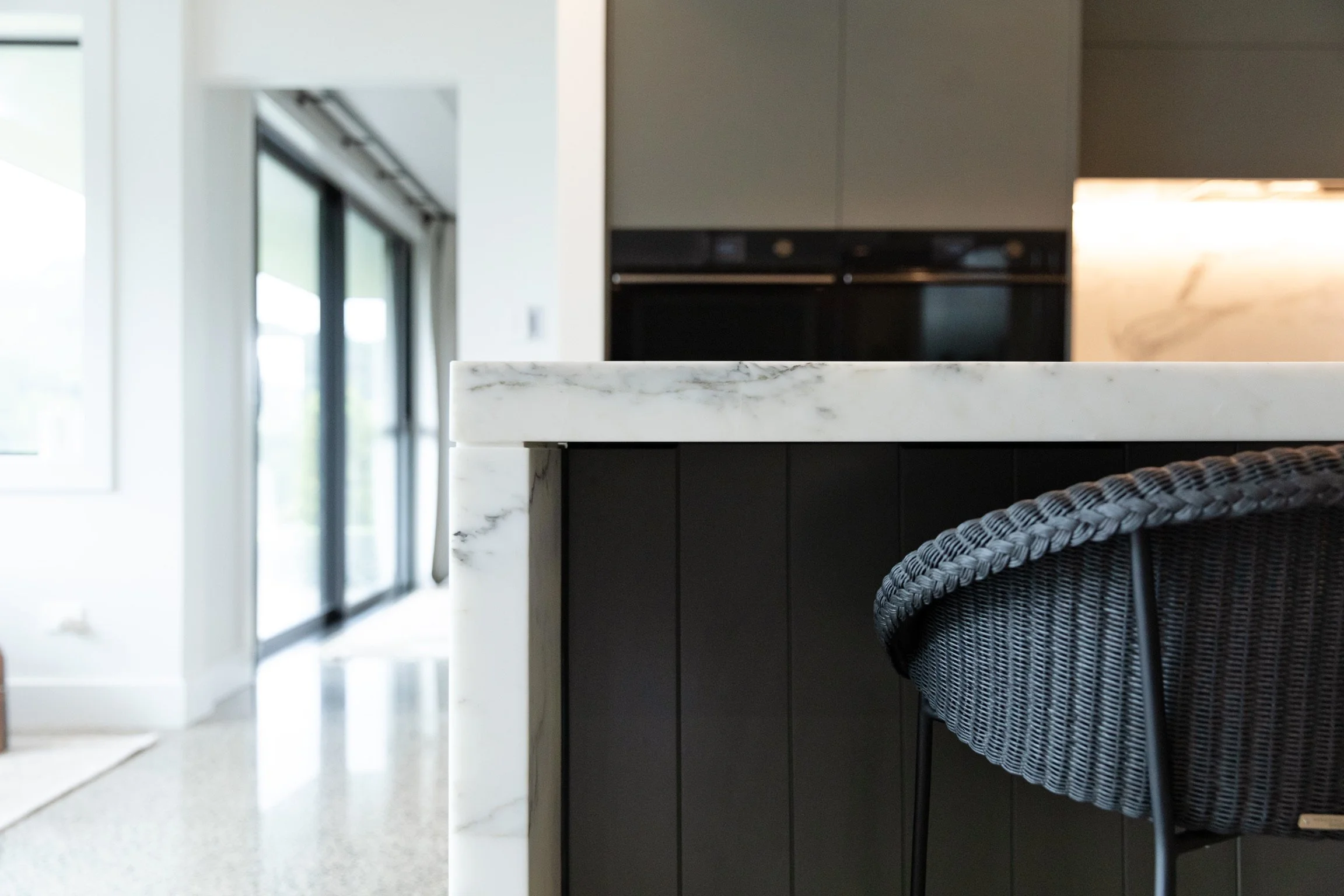 Close-up of a white marble countertop in a modern kitchen, with a black woven chair partially visible.