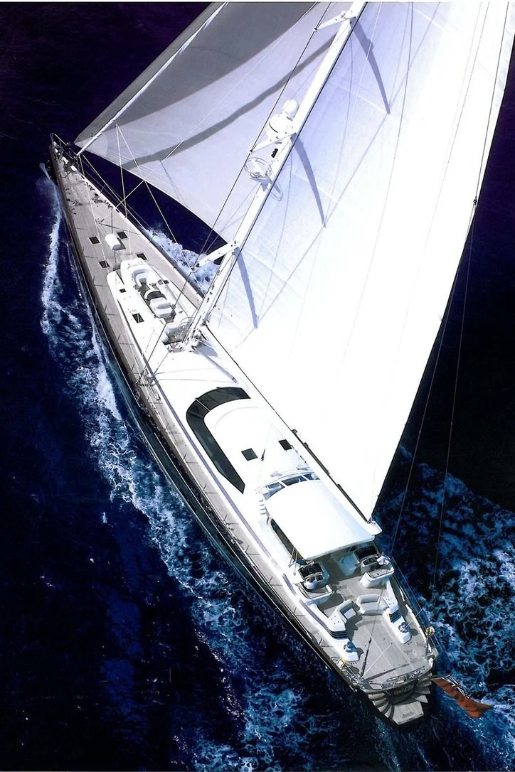 Aerial view of a large white sailing yacht with sails up, moving through deep blue ocean water.