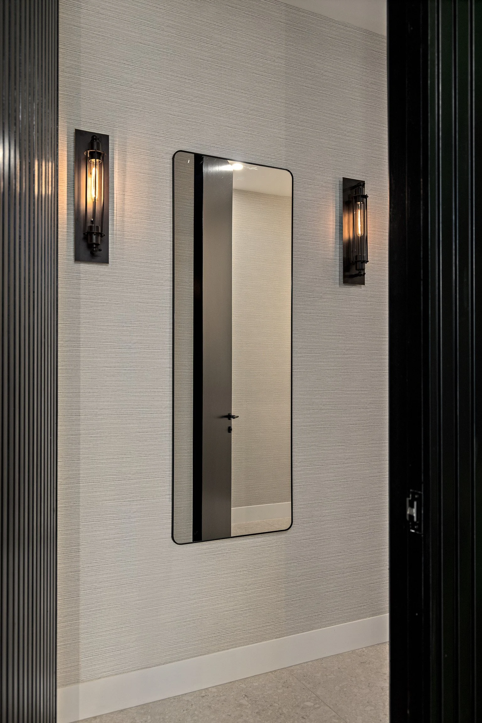 A rectangular mirror mounted on a beige textured wall, with two black wall sconces with exposed filament bulbs on either side, beneath ceiling lighting, near a corner of a room with a black vertical slatted wall to the right.