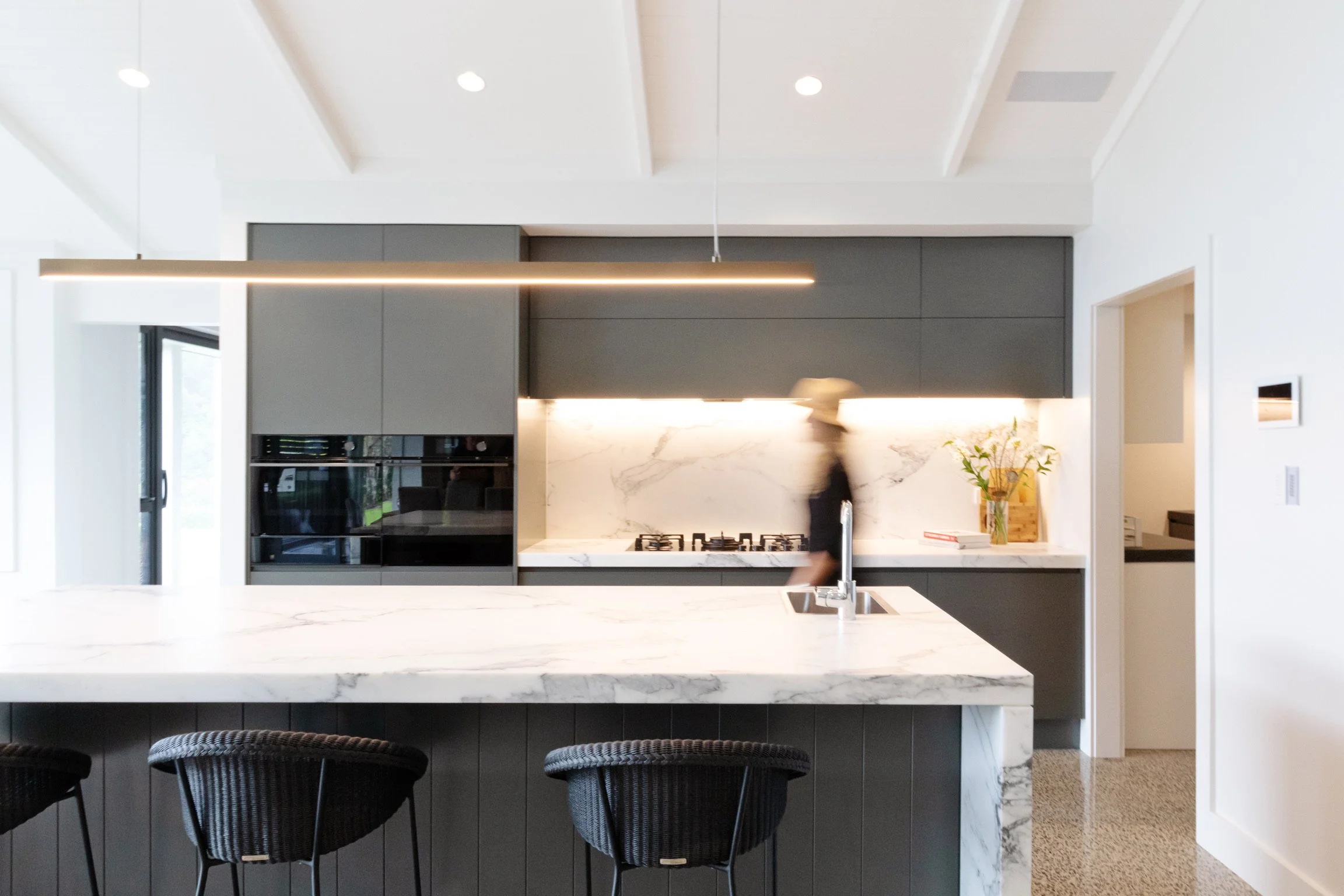 Modern kitchen with marble island, gray cabinets, built-in oven, gas stove, and black chairs in the foreground.